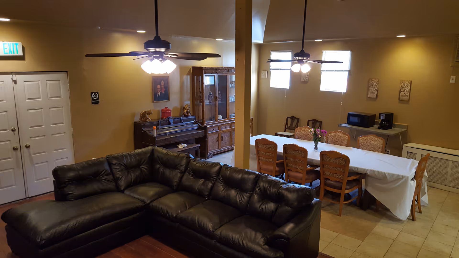 Interior view of a senior living facility common area with a black leather sectional sofa in the foreground, a dining table with chairs covered with a white tablecloth and a vase of flowers, two ceiling fans with lights, a piano against the wall, a wooden cabinet, a microwave and coffee maker on a small table, and beige walls with framed artwork.