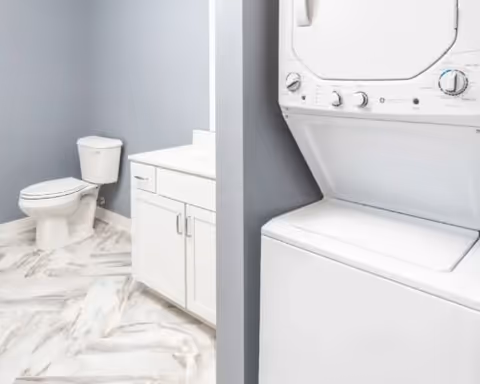 Small bathroom with a white vanity and toilet, a stacked washer-dryer unit, gray walls, and marble-look flooring.