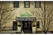 Entrance of Terence Cardinal Cooke Health Care Center with a green dome canopy above the doorway, flanked by leafless trees and beige building walls with windows.