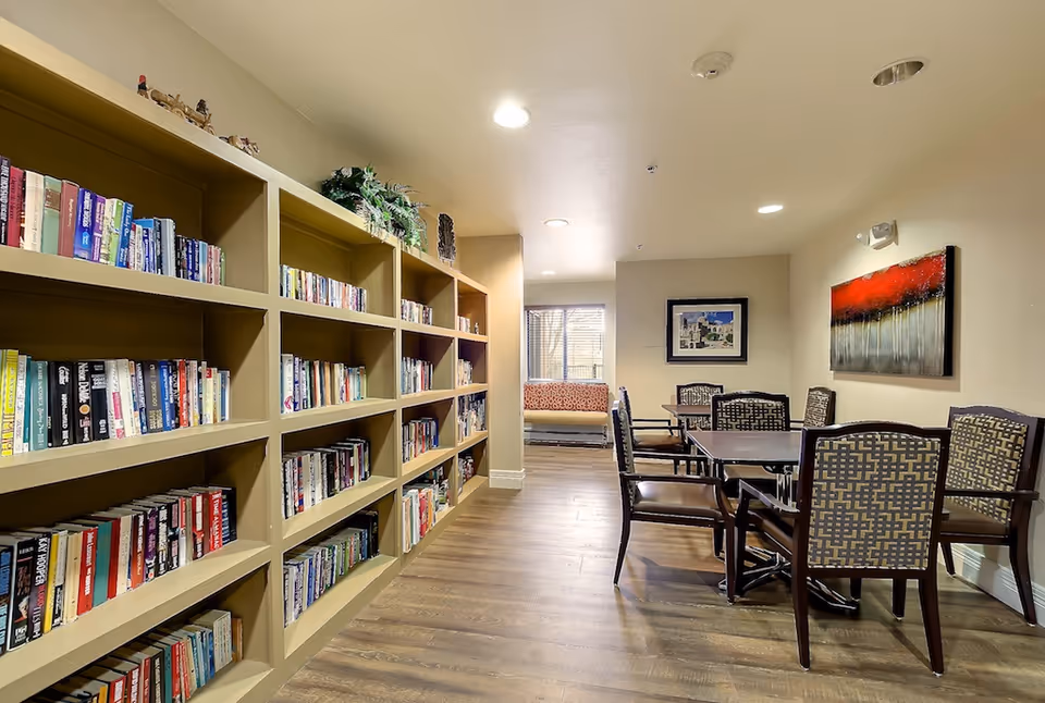 Interior view of a senior living facility room with a long bookshelf filled with books on the left side, a wooden floor, and a seating area with a table and six chairs on the right. The room has beige walls, two framed artworks on the walls, and a window with a cushioned bench underneath at the far end.