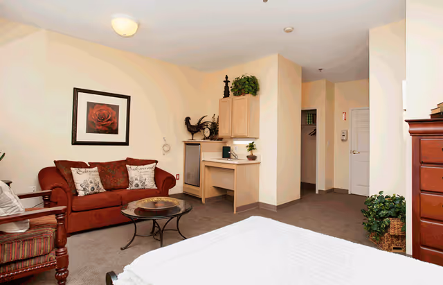 A cozy living space featuring a red sofa with decorative pillows, a glass coffee table, a wooden armchair with striped cushions, a built-in desk with cabinets, and a bed partially visible in the foreground. The walls are light-colored, and there is a framed picture of a red rose above the sofa. The room also includes some plants and decorative items.