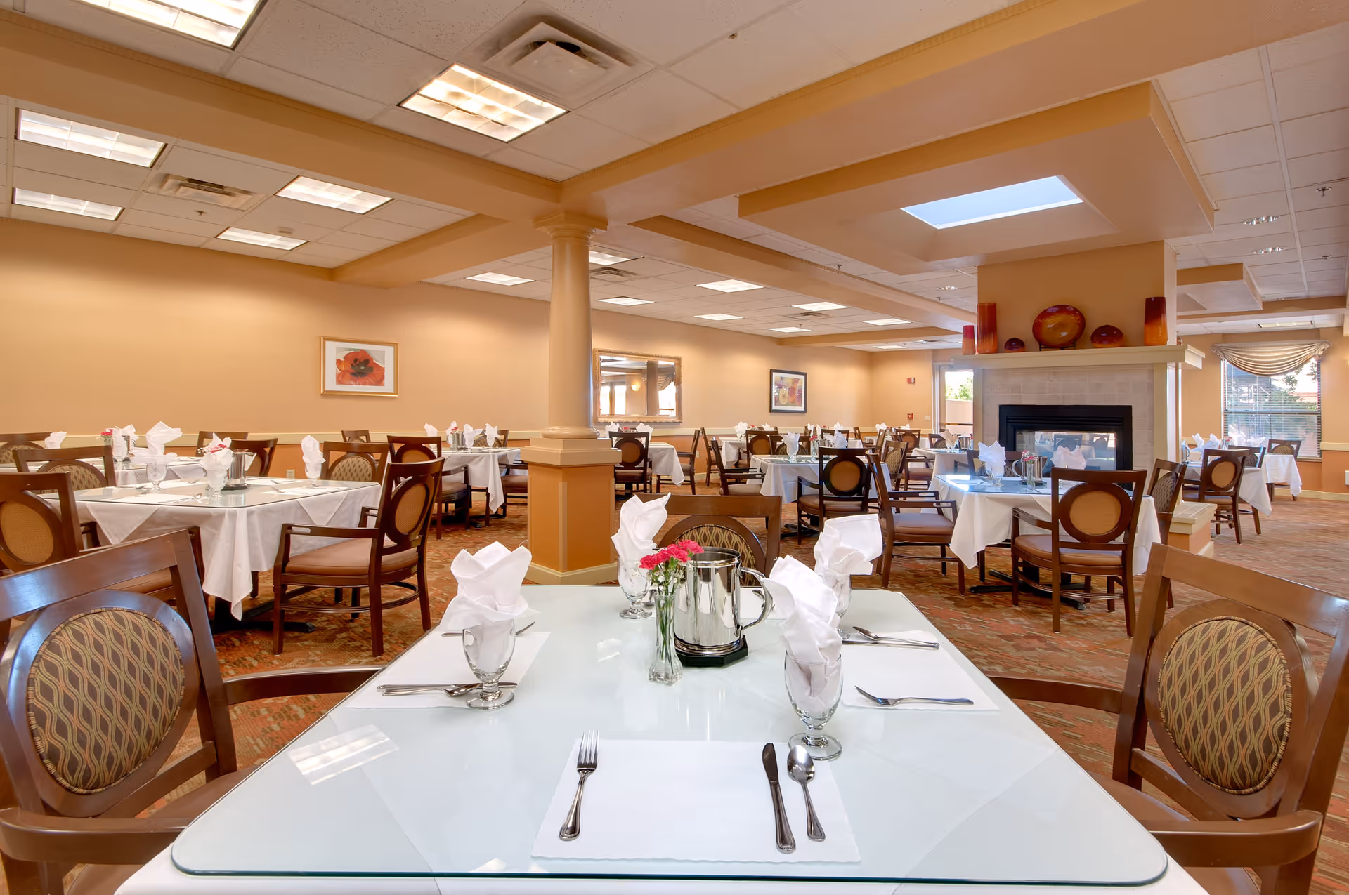 A spacious dining room with multiple tables covered in white tablecloths, each set with folded napkins, glassware, and silverware. The room features warm beige walls, a carpeted floor, a fireplace with decorative items on the mantle, and large windows allowing natural light to enter.
