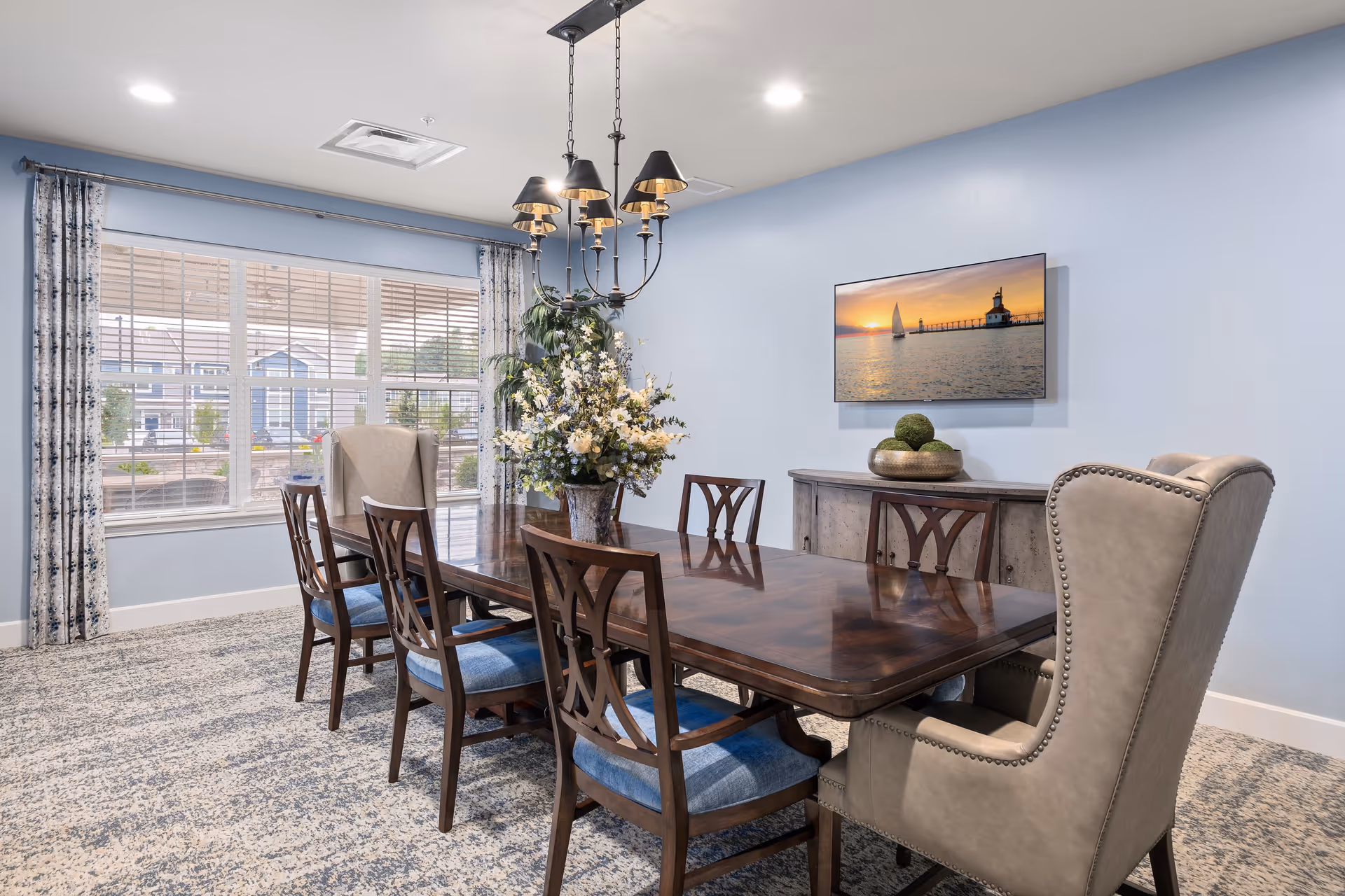 A dining room with a long wooden table surrounded by six chairs, two of which are upholstered armchairs. A large floral arrangement is placed in the center of the table. The room has light blue walls, a patterned carpet, and a window with blinds and curtains. A chandelier with black lampshades hangs above the table, and a flat-screen TV displaying a sunset over water is mounted on the wall above a wooden sideboard with decorative items.