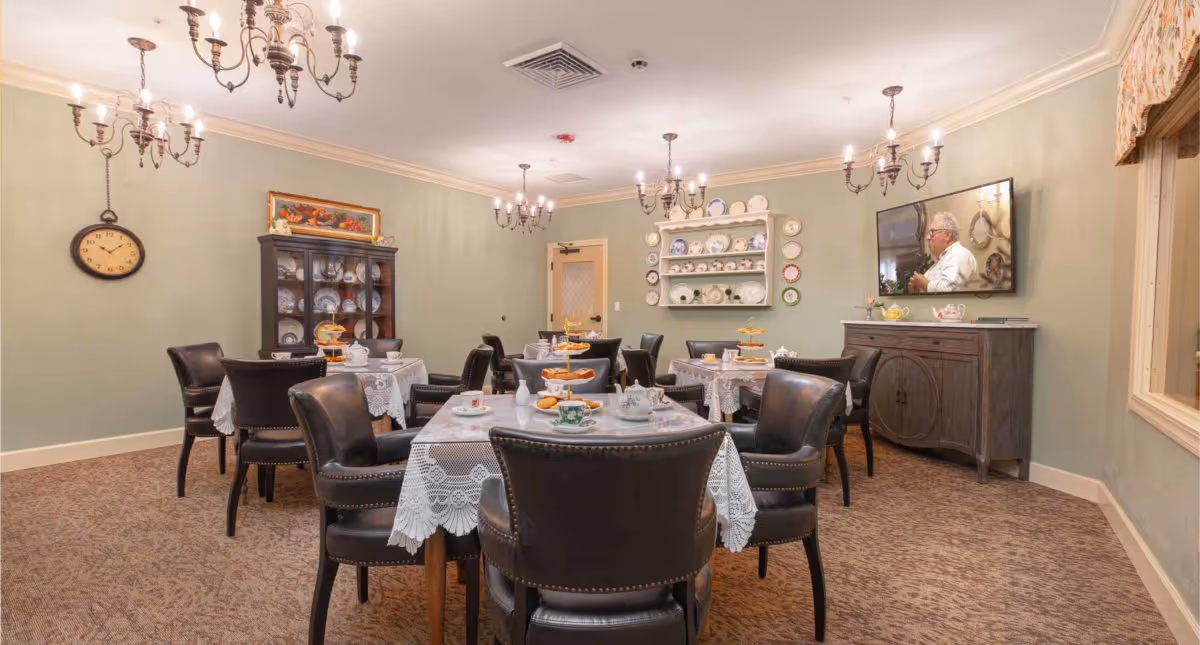 Communal dining room with round tables set with lace tablecloths and tea service, leather chairs, chandeliers, a china cabinet and a wall-mounted TV.
