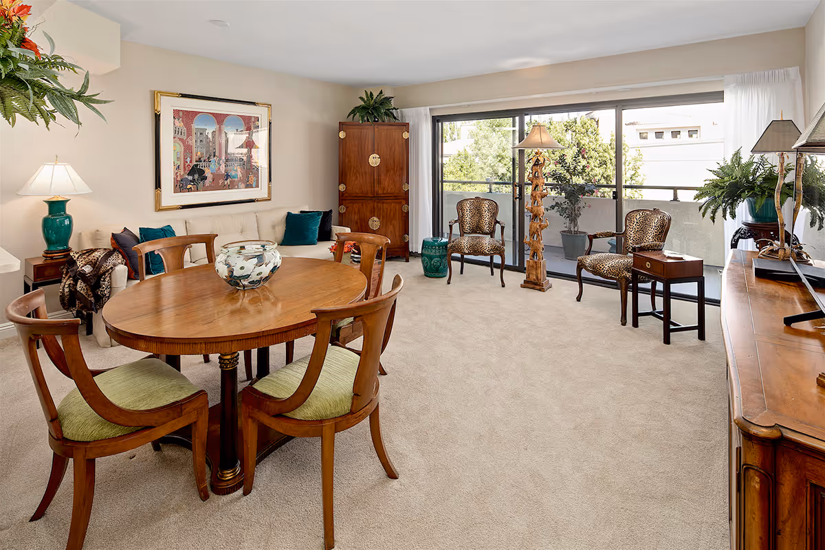 A bright living room with beige carpet and walls, featuring a wooden oval dining table with four chairs that have green cushions. There is a cream-colored sofa with teal and brown pillows, a wooden cabinet, and two leopard print armchairs near sliding glass doors leading to a balcony with plants. The room is decorated with various lamps, plants, and a framed artwork on the wall.