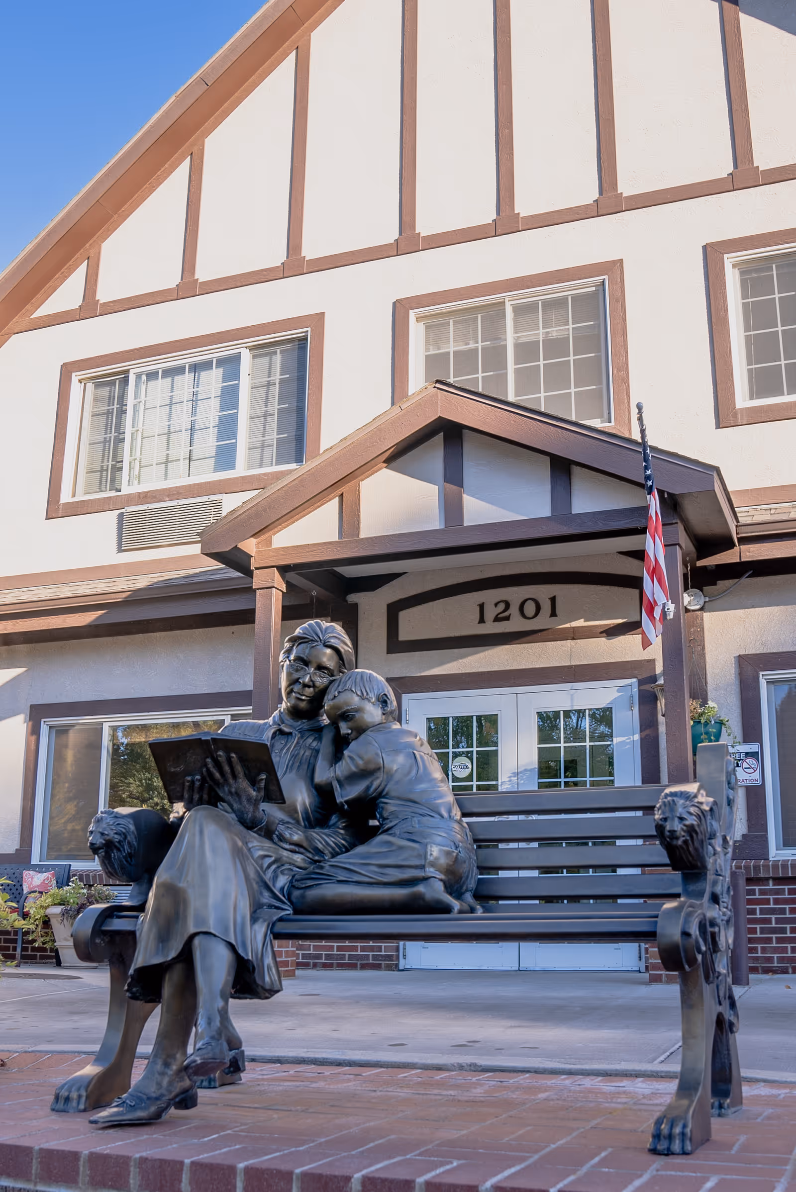 Bronze statue of a woman reading a book to a child sitting on a decorative bench in front of a building with the address number 1201 and an American flag.