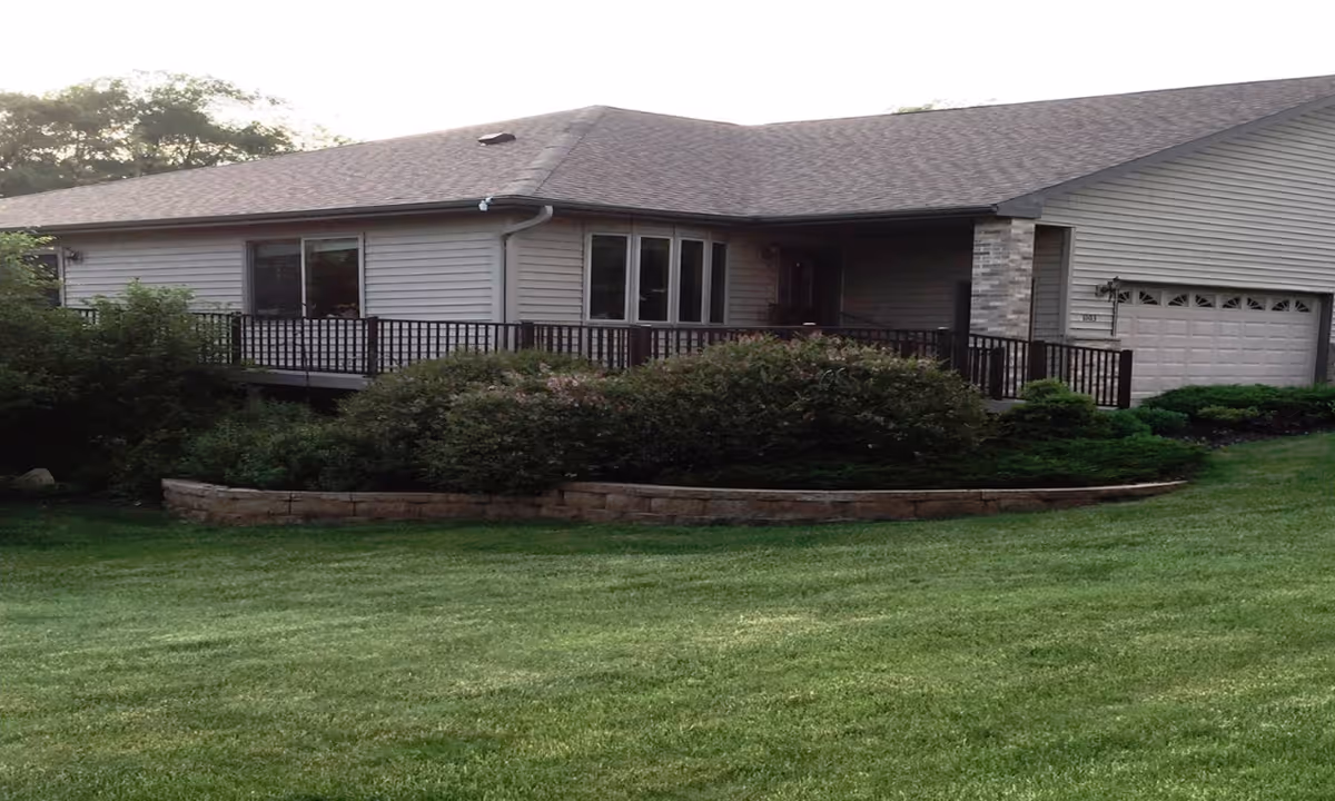 Single-story building with a front deck and attached garage set behind a manicured lawn and shrubs.