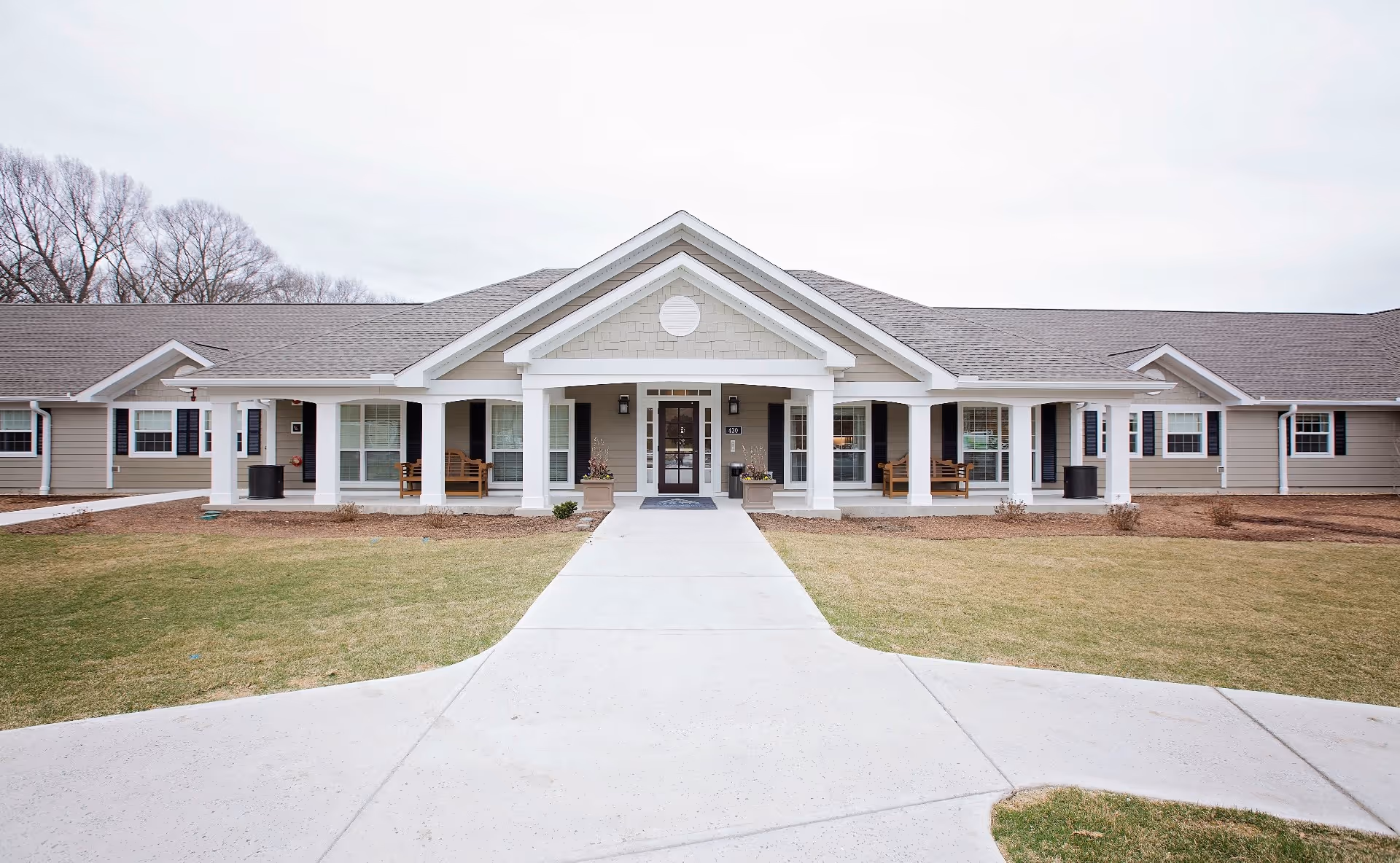 Front exterior view of a single-story senior living facility building with a covered entrance supported by white columns, beige siding, multiple windows with black shutters, and a concrete walkway leading to the entrance. There are benches on the porch and a grassy lawn in front.