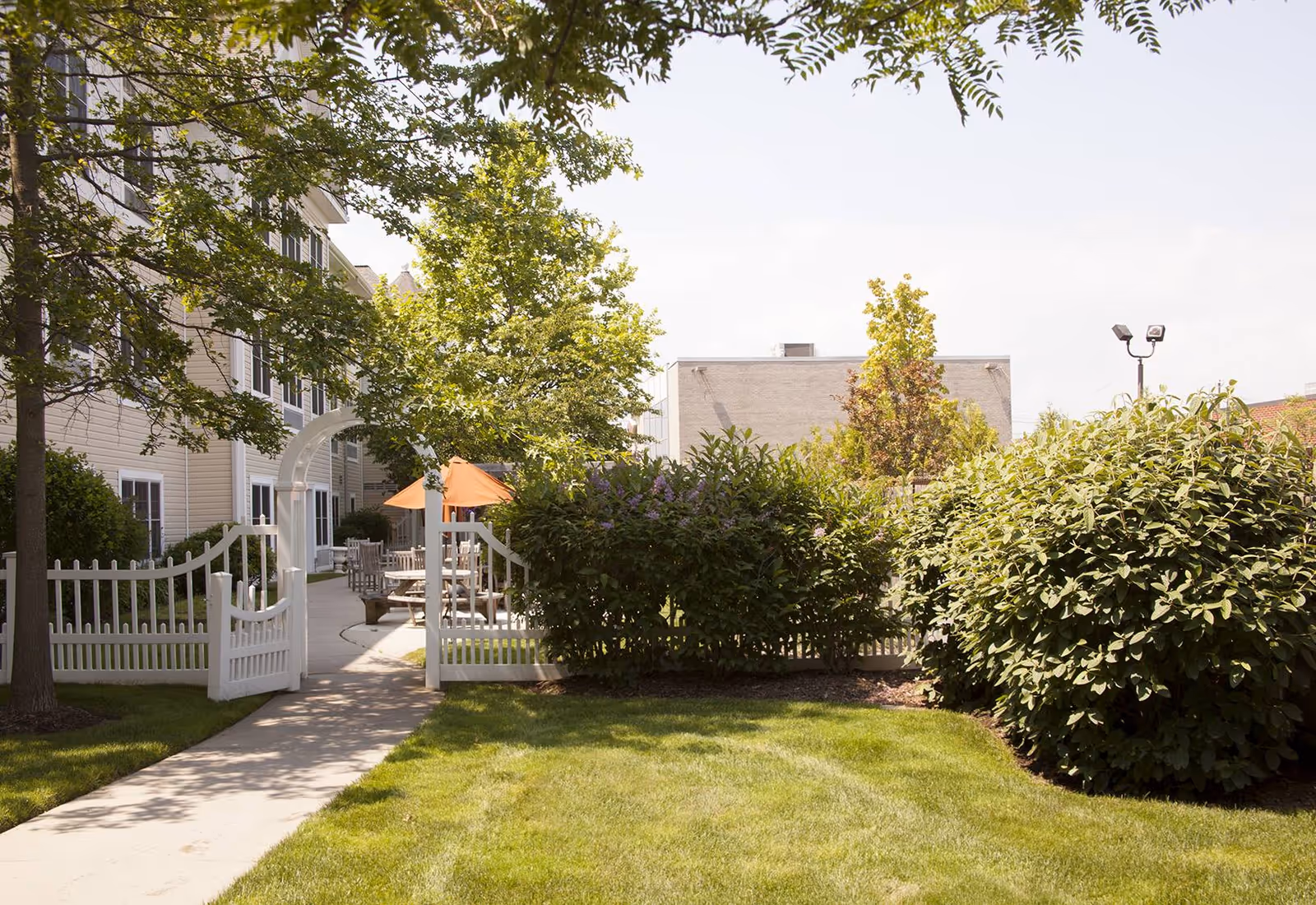 A sunny outdoor garden area at Sunrise of West Babylon featuring a white picket fence gate, green grass, trees, bushes, and a patio with tables and chairs shaded by orange umbrellas.