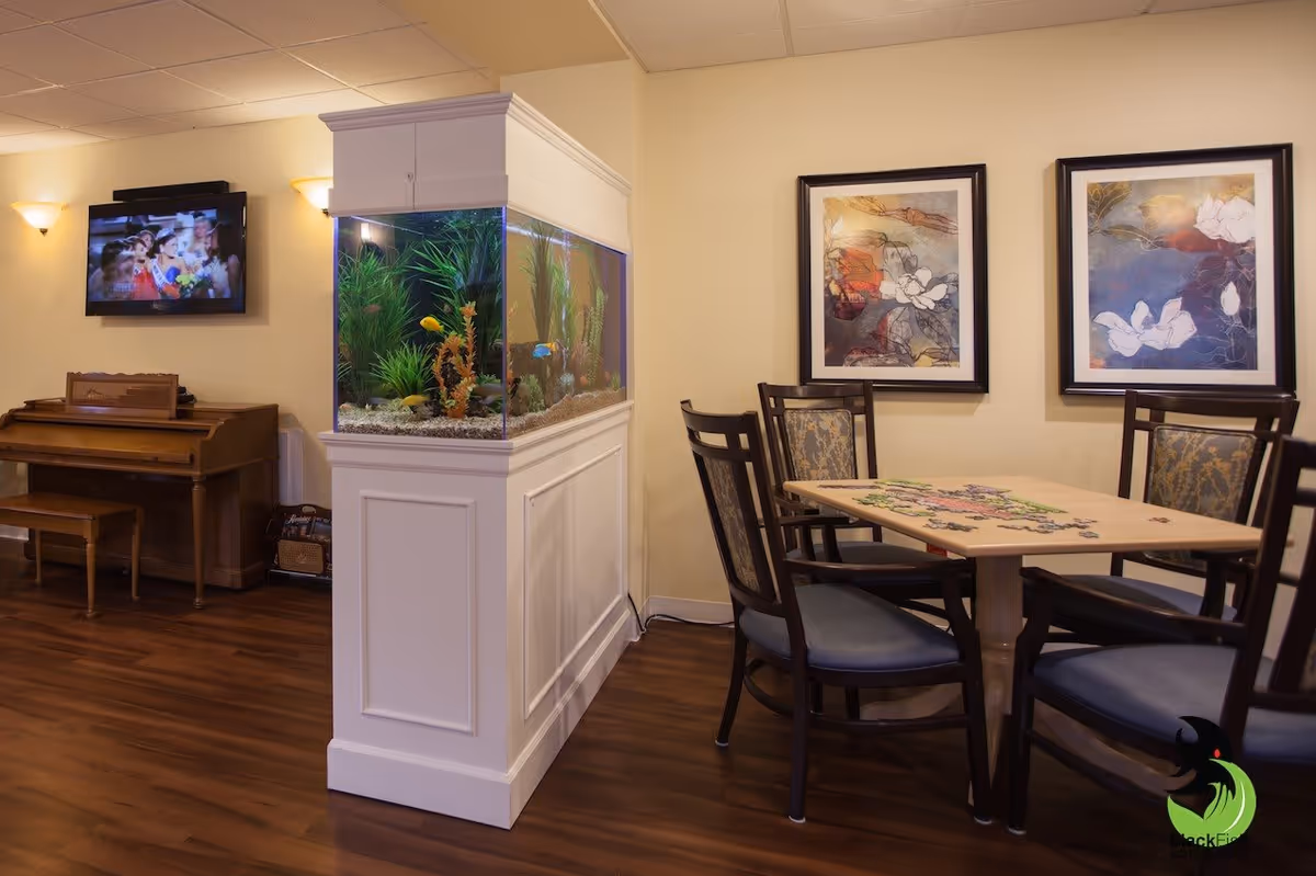 Communal lounge with a large built-in aquarium, table and chairs, a piano, and framed artwork on the walls.