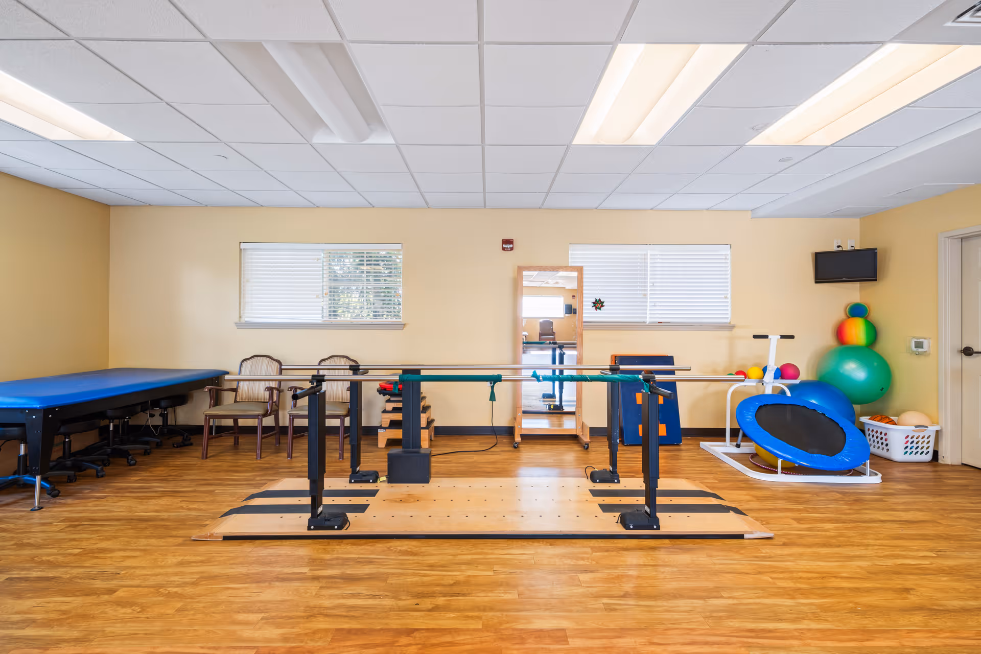 A physical therapy or rehabilitation room with parallel bars for walking exercises in the center, a blue therapy table on the left, two chairs against the wall, a full-length mirror, and various exercise equipment including a small trampoline, exercise balls, and a basket of therapy tools on the right. The room has wood flooring, beige walls, and two windows with blinds.