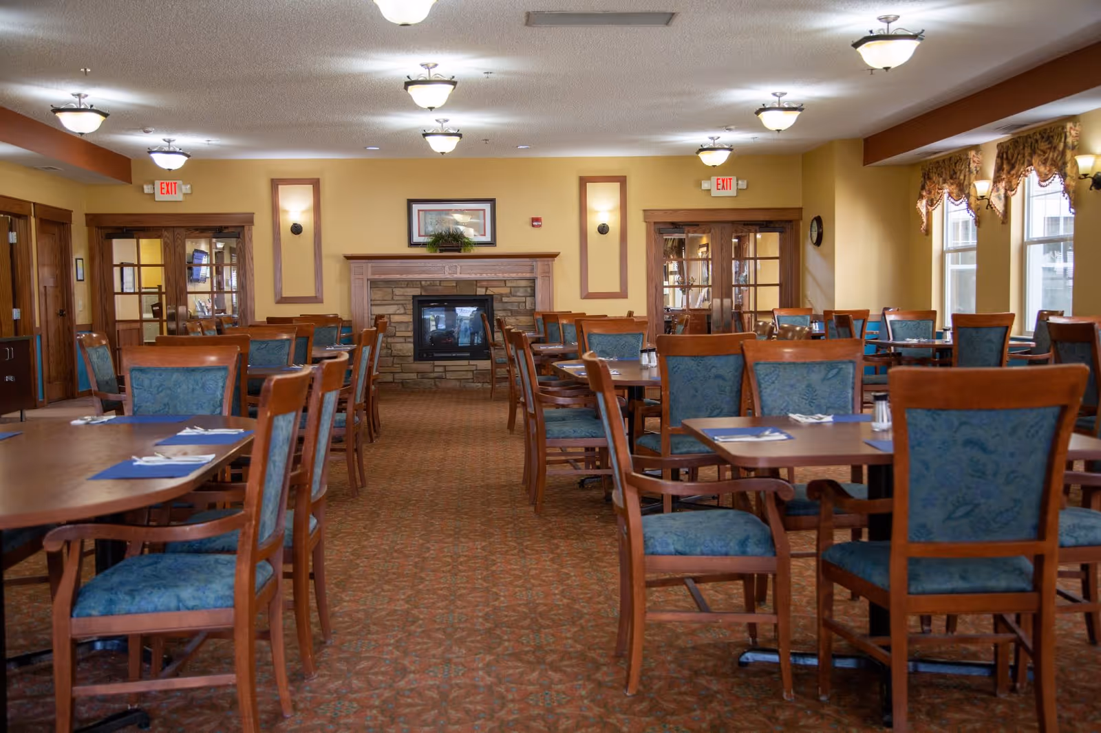 A spacious dining room with multiple wooden tables and chairs upholstered in blue fabric. The room features a carpeted floor, a stone fireplace with a wooden mantel, framed artwork above the fireplace, and several ceiling lights. Large windows with decorative curtains allow natural light to enter the room.