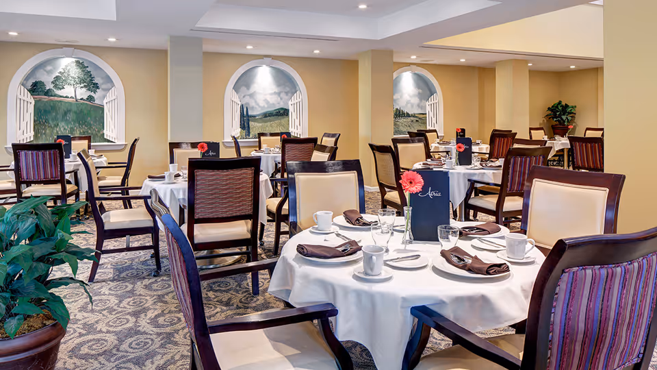 A dining room in a senior living facility with multiple round tables covered with white tablecloths. Each table is set with cups, glasses, plates, and brown napkins. There are wooden chairs with cushioned seats around the tables. The walls have painted murals depicting outdoor scenes with trees and fields, framed by arched window-like designs. A potted plant is visible in the foreground.
