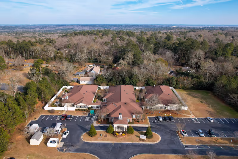 Aerial view of PruittPlace - Wesleyan senior living facility surrounded by trees and open land, showing a large building with multiple wings, a parking lot with several cars, and a circular driveway at the entrance.