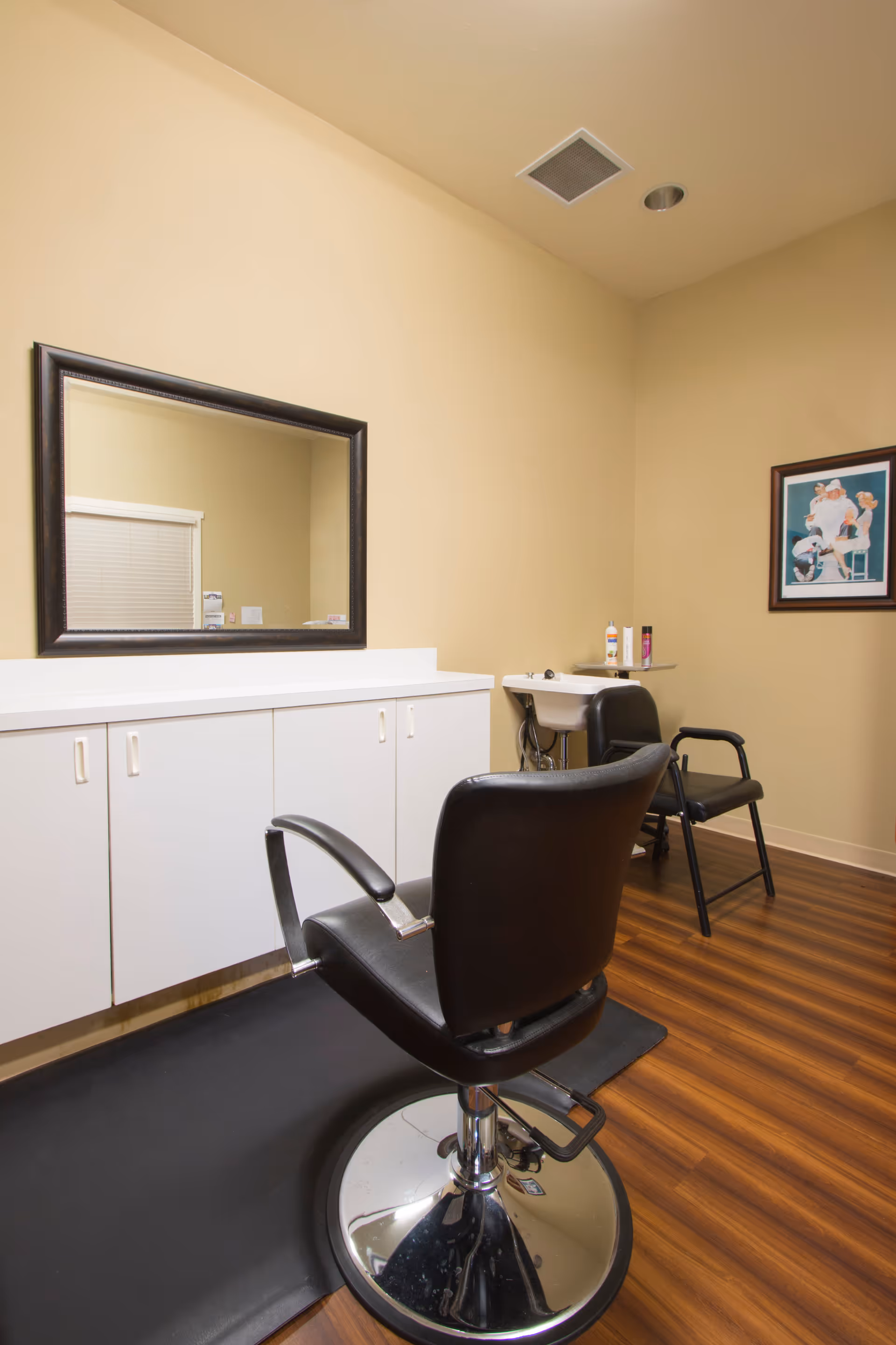 Interior of a small salon area with a black salon chair on a black mat in front of a large mirror mounted above white cabinets. In the background, there is a small white sink with hair care products on a shelf and a black chair next to it. The room has beige walls and wood flooring, with a framed picture hanging on the wall.