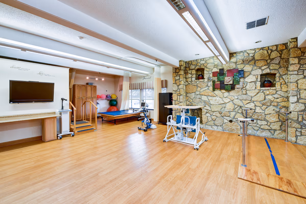 A spacious therapy or rehabilitation room with wooden flooring, a stone accent wall, and various exercise equipment including a stationary bike, parallel bars, and therapy tables. The room has large windows with curtains allowing natural light, a wall-mounted TV, and motivational quotes on the walls.