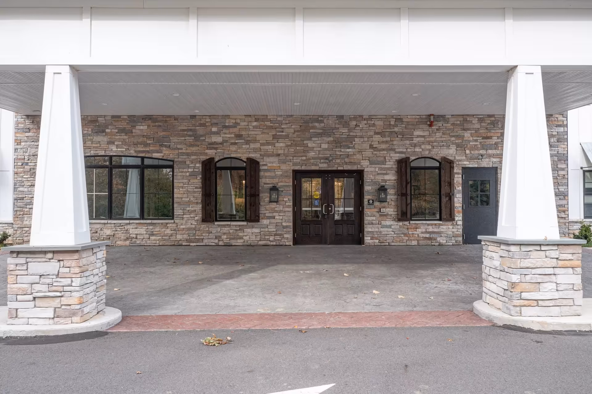 Covered entrance area of a building with stone walls, two large white pillars, three windows with dark wooden shutters, and a double glass door in the center.