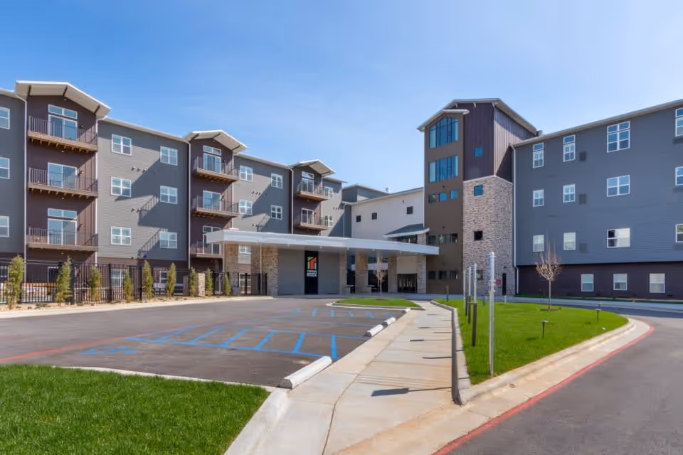 Exterior view of Turners Rock senior living facility showing a modern multi-story building with balconies, large windows, a covered entrance, and a parking area with handicap spaces under a clear blue sky.