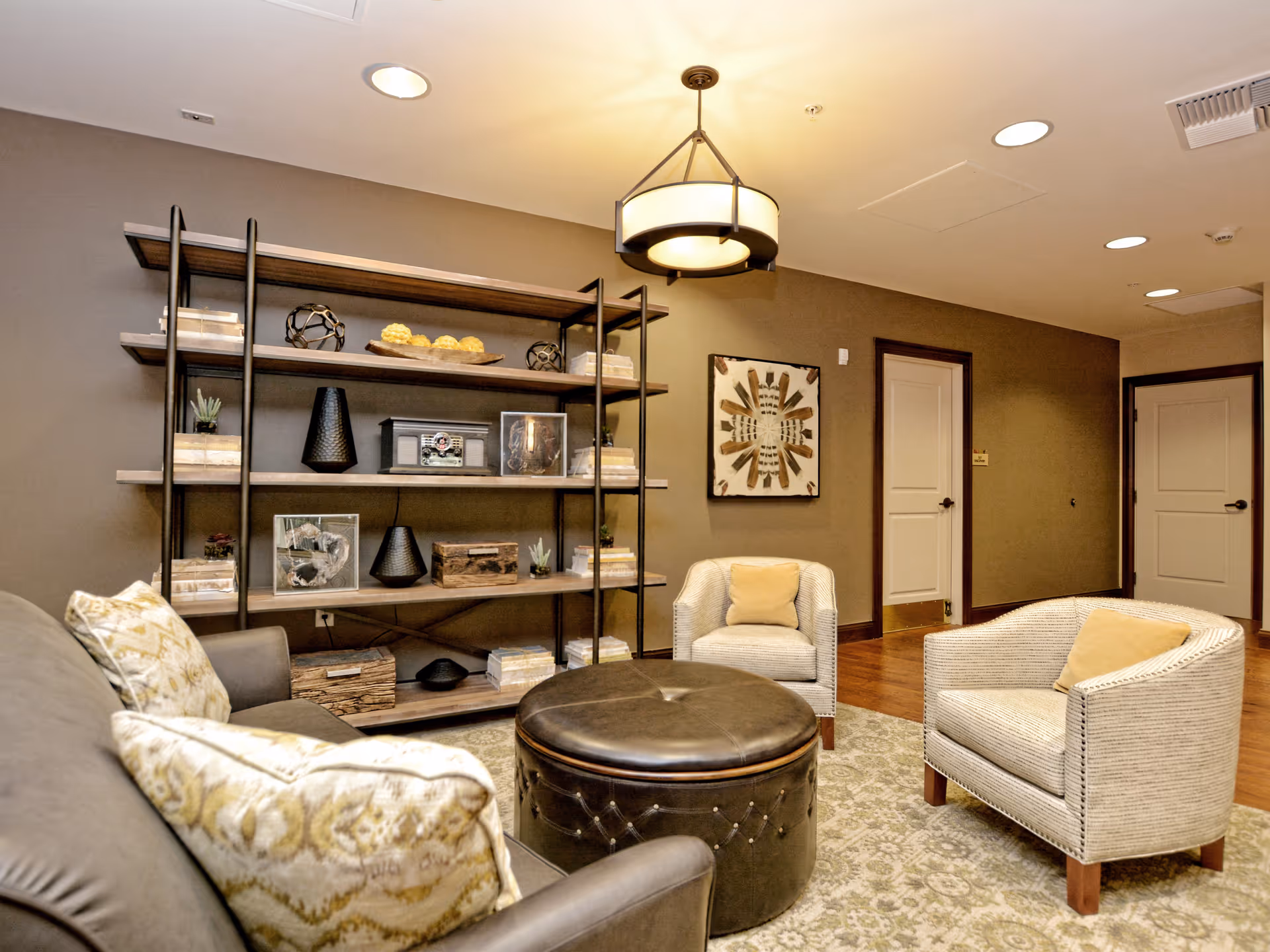 Cozy sitting area with armchairs, a round leather ottoman, decorative shelving, and warm lighting.