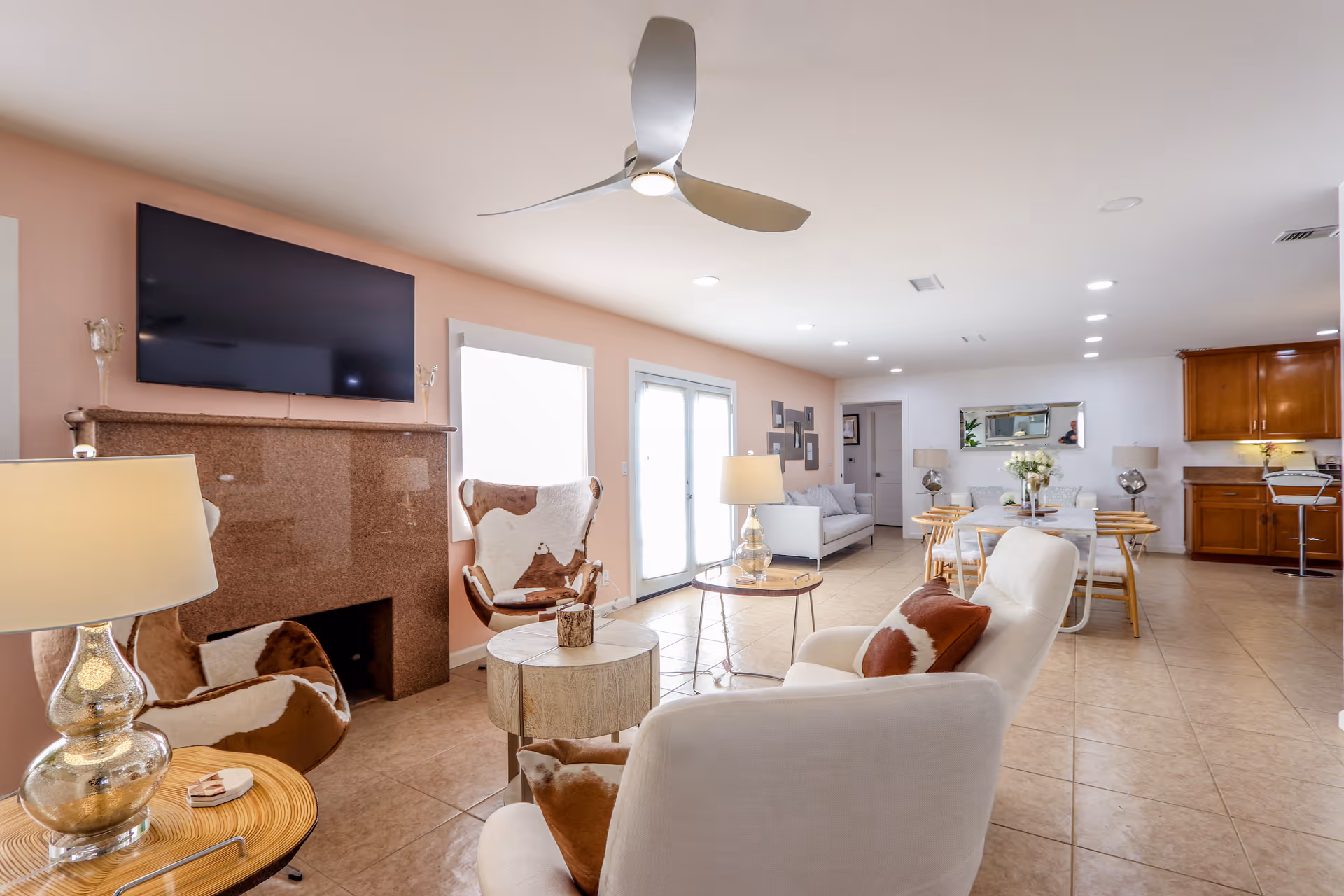 A bright and spacious living room with a mounted flat-screen TV above a brown fireplace, two armchairs with cowhide upholstery, a white armchair with a brown pillow, a round wooden side table with a lamp, and a ceiling fan. In the background, there is a dining area with a table and chairs, a large mirror on the wall, and a kitchen with wooden cabinets.