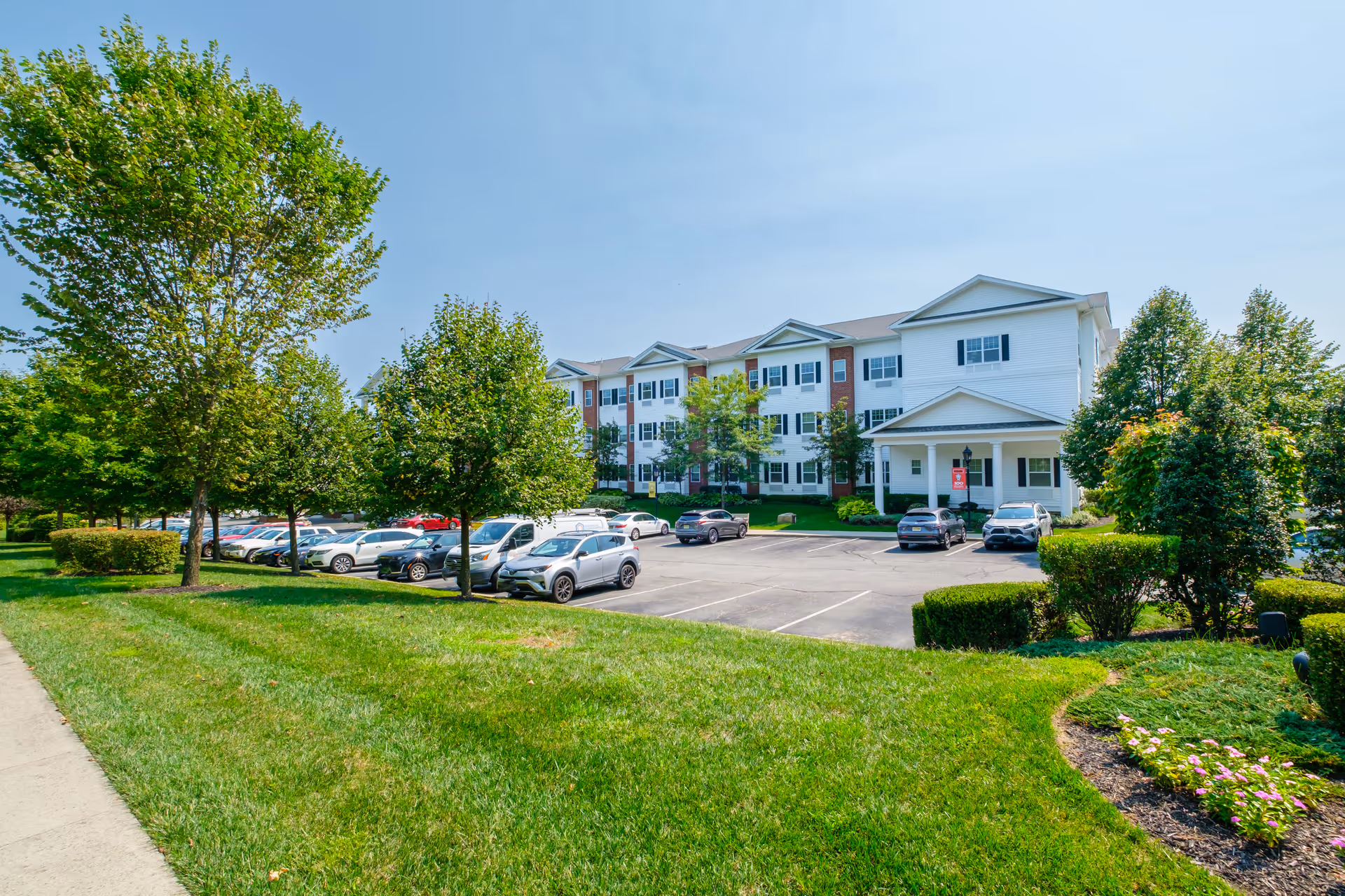 Exterior view of a senior living facility building with white siding and brick accents, surrounded by green trees and a well-maintained lawn. There is a parking lot with several cars parked in front of the building under a clear blue sky.
