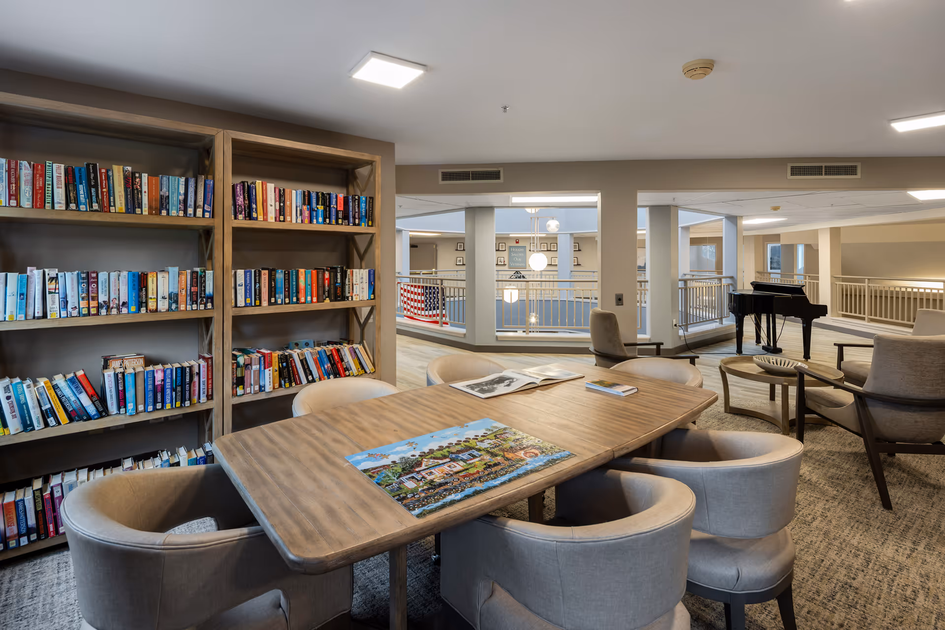 A cozy senior living community common area with a wooden table surrounded by cushioned chairs. On the table, there is a partially completed jigsaw puzzle and an open book. Behind the table, there are bookshelves filled with books. In the background, there is a black grand piano and additional seating areas with armchairs. The space is well-lit with ceiling lights and has a neutral color palette with beige walls and carpeted floors.