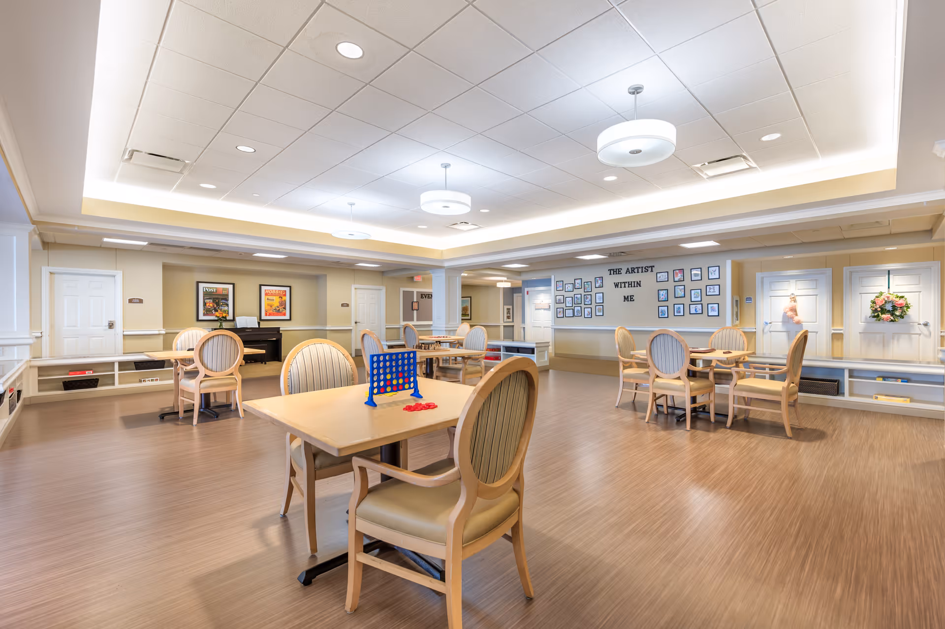A spacious, well-lit activity room with several tables and chairs arranged for games and socializing. One table has a Connect Four game set up. The walls are decorated with framed pictures and a display titled 'The Artist Within Me' showcasing various artworks. The room has a light wood floor and a white ceiling with recessed and hanging lights.