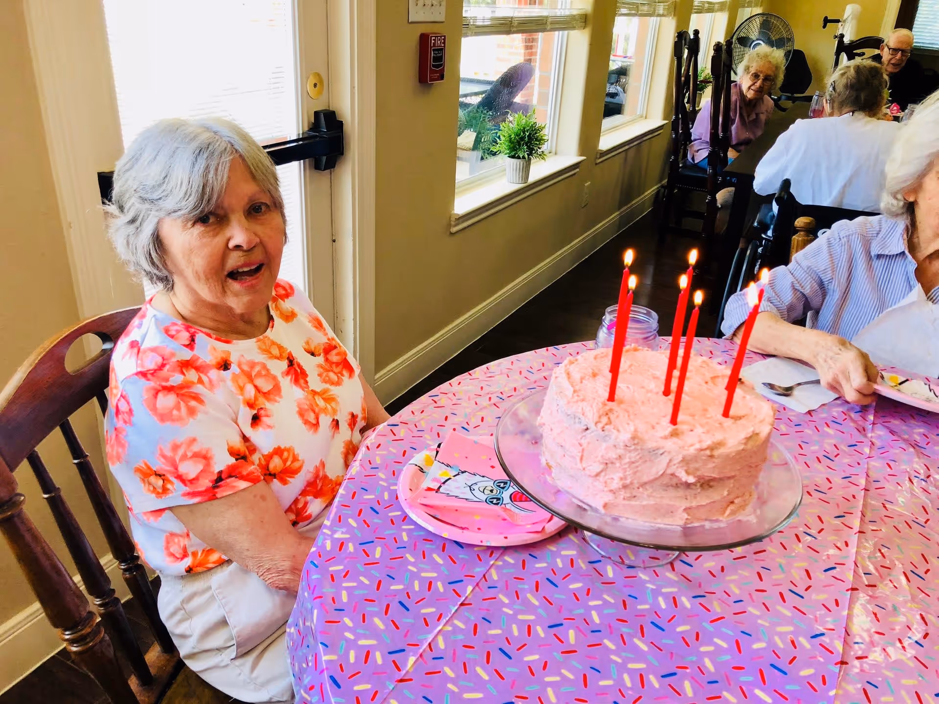 Elderly residents seated around a decorated table with a pink frosted birthday cake and lit candles in a brightly lit dining area.
