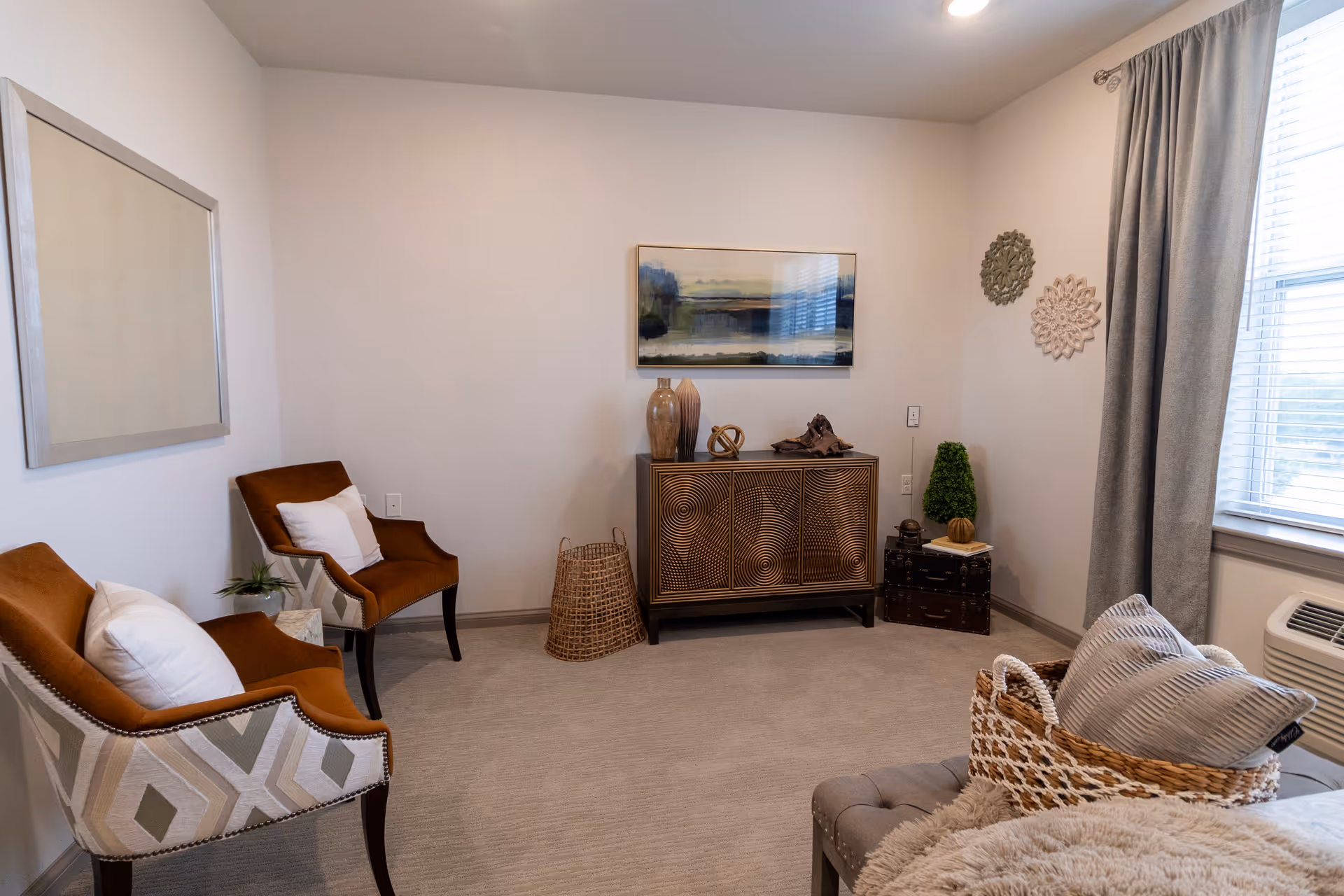 A bright, neutral sitting room with two upholstered chairs, a decorative cabinet, and a window with curtains.