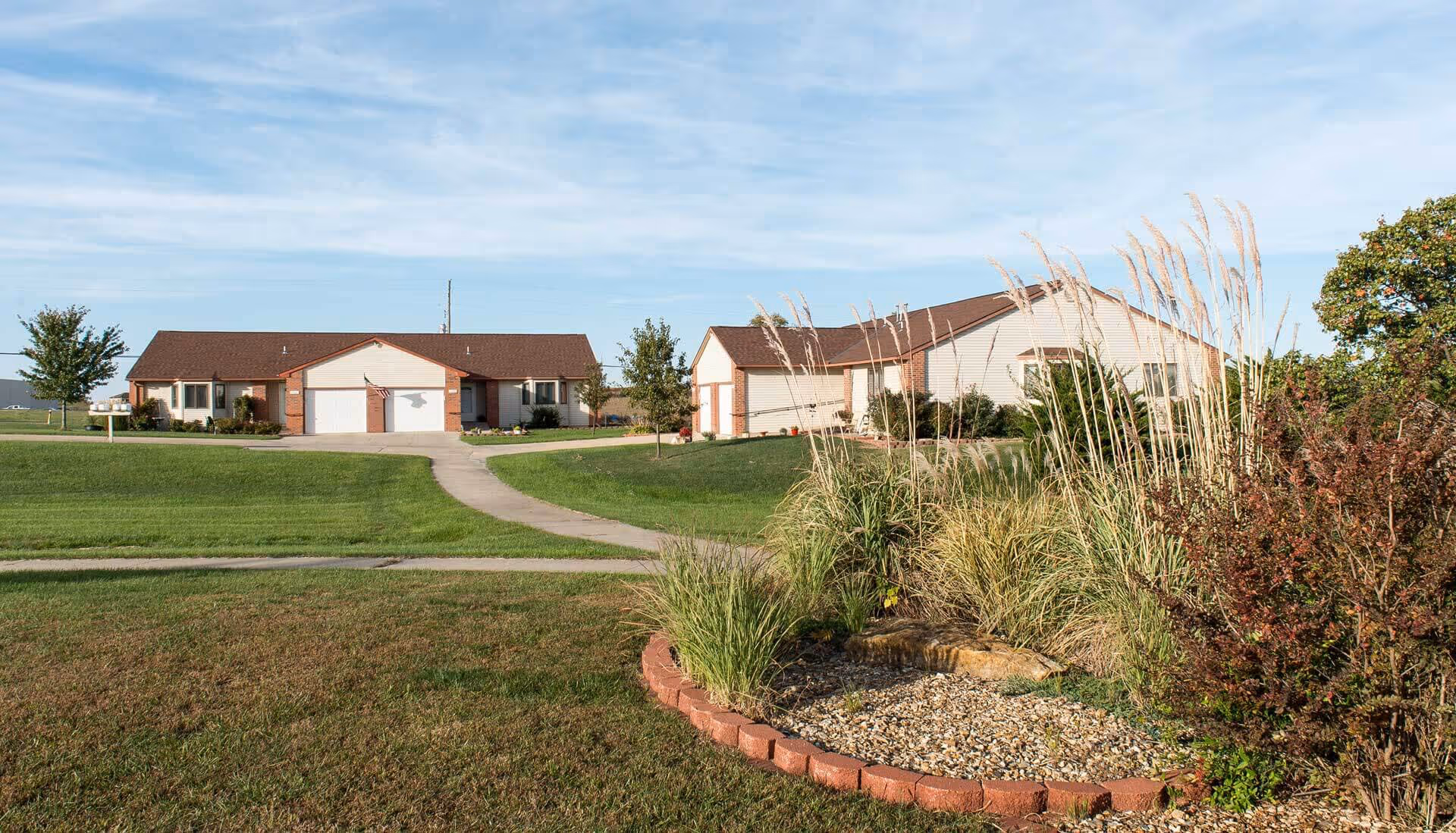 View of a senior living facility with two single-story buildings featuring garages, surrounded by well-maintained green lawns and landscaped garden beds with ornamental grasses and shrubs under a clear blue sky.