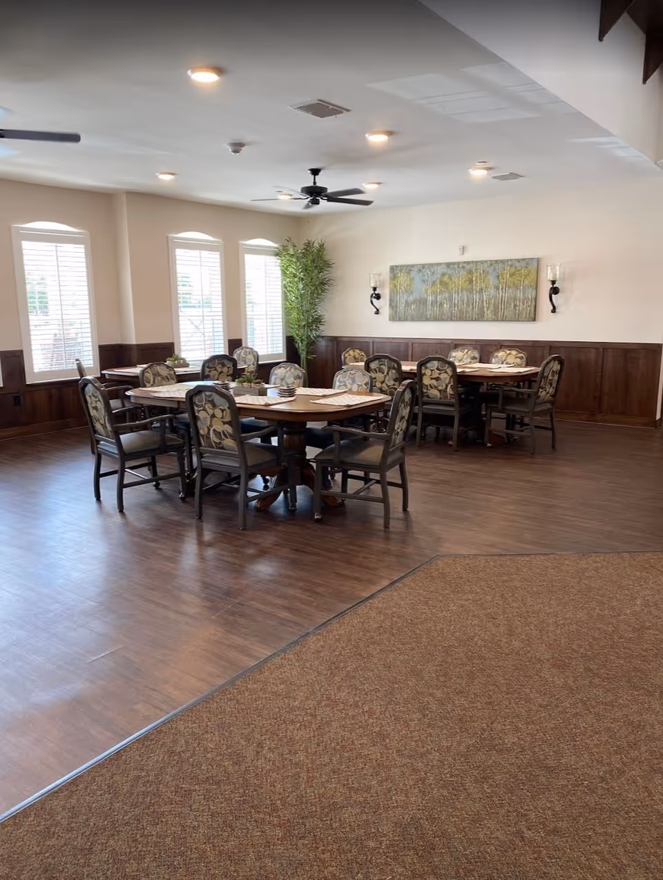 A dining room with two round wooden tables surrounded by cushioned chairs with patterned upholstery. The room has large windows with white shutters, a ceiling fan, recessed lighting, wood paneling on the walls, a large potted plant in the corner, and a landscape painting on the wall.