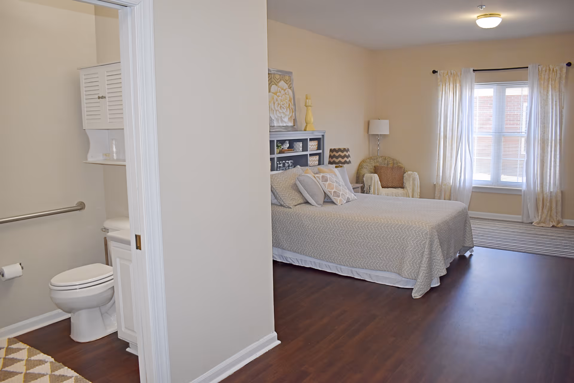 A bedroom with a bed covered in a light gray patterned bedspread and several pillows. There is a wicker chair with a cushion and a floor lamp near a window with sheer curtains. To the left, a bathroom is partially visible with a toilet, a grab bar, and a cabinet above the toilet. The room has wooden flooring and beige walls.