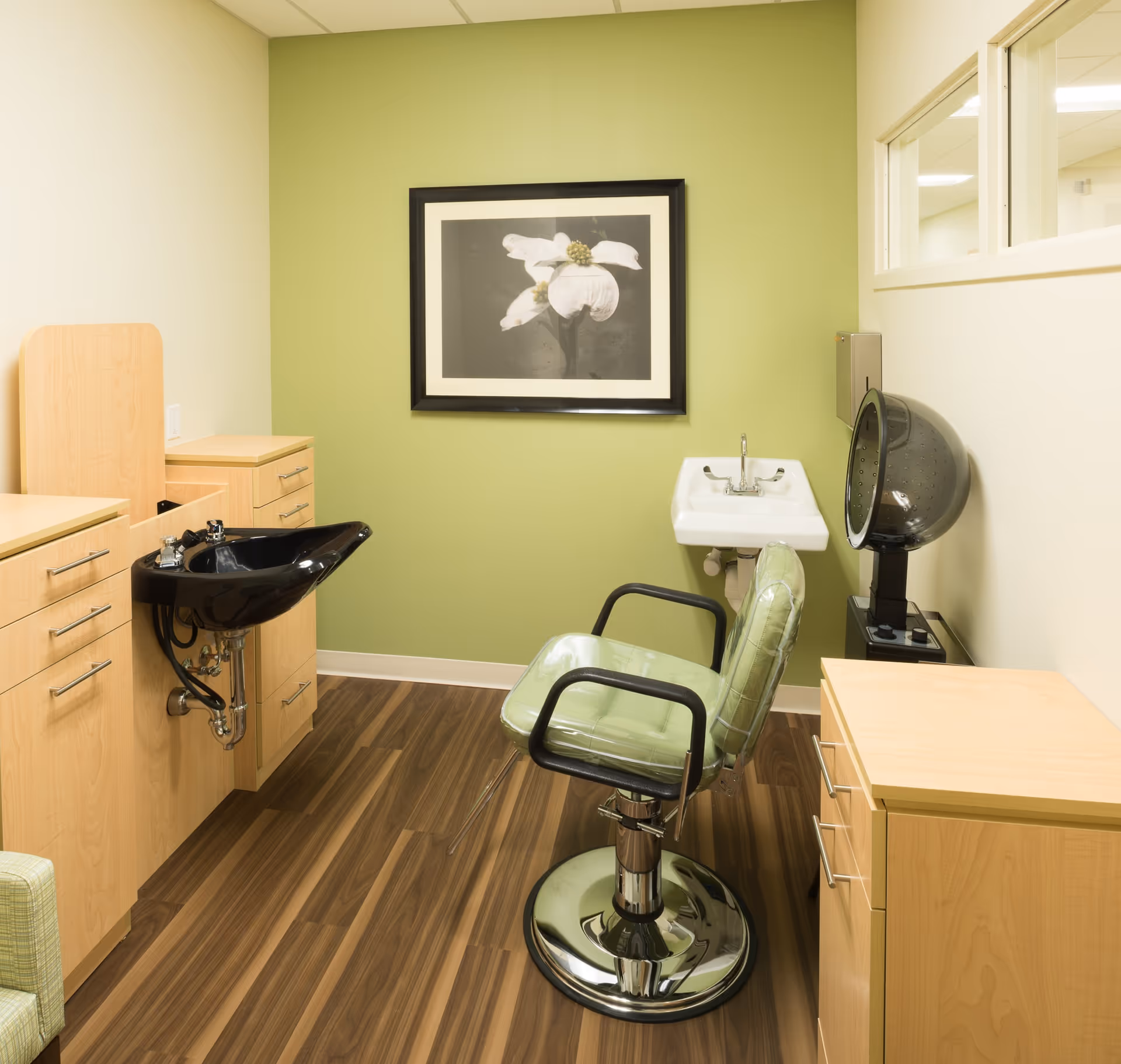 A small salon room with a green salon chair, a black hair washing sink, a white wall-mounted sink, wooden cabinets, and a black hair dryer. The walls are painted light green and beige, and there is a framed picture of a white flower on the green wall.