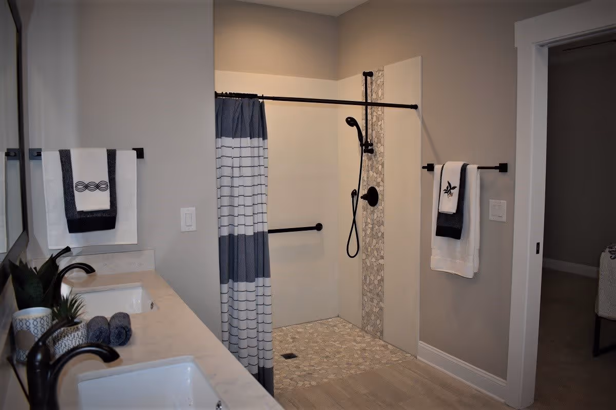 Modern bathroom with a walk-in shower featuring a pebble tile floor and a vertical pebble tile accent strip. The shower has a black handheld showerhead and a black grab bar. There is a blue and white striped shower curtain on a black rod. To the left, a double sink vanity with black faucets, a potted plant, and neatly folded towels is visible. Two towel racks hold white and dark blue towels with decorative embroidery. The bathroom walls are painted light gray, and there is an open doorway leading to another room.