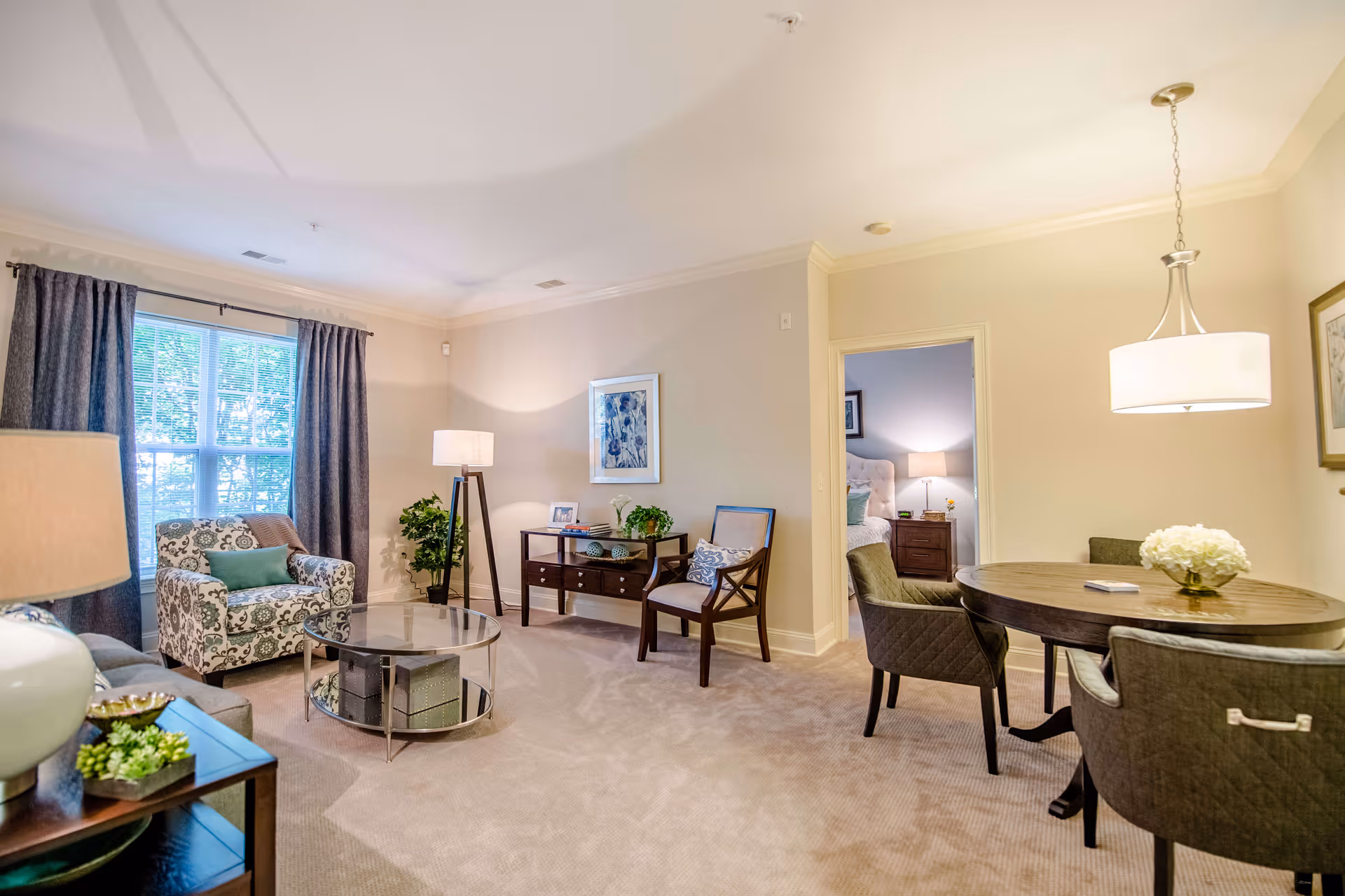 A well-lit living room and dining area in a senior living facility. The living room features a patterned armchair, a glass coffee table, a wooden side table with a lamp and plants, and a floor lamp. The dining area has a round wooden table with four upholstered chairs and a hanging light fixture above. A doorway leads to a bedroom with a bed, nightstand, and lamp visible. The room has beige walls, carpeted floors, and a large window with dark curtains.