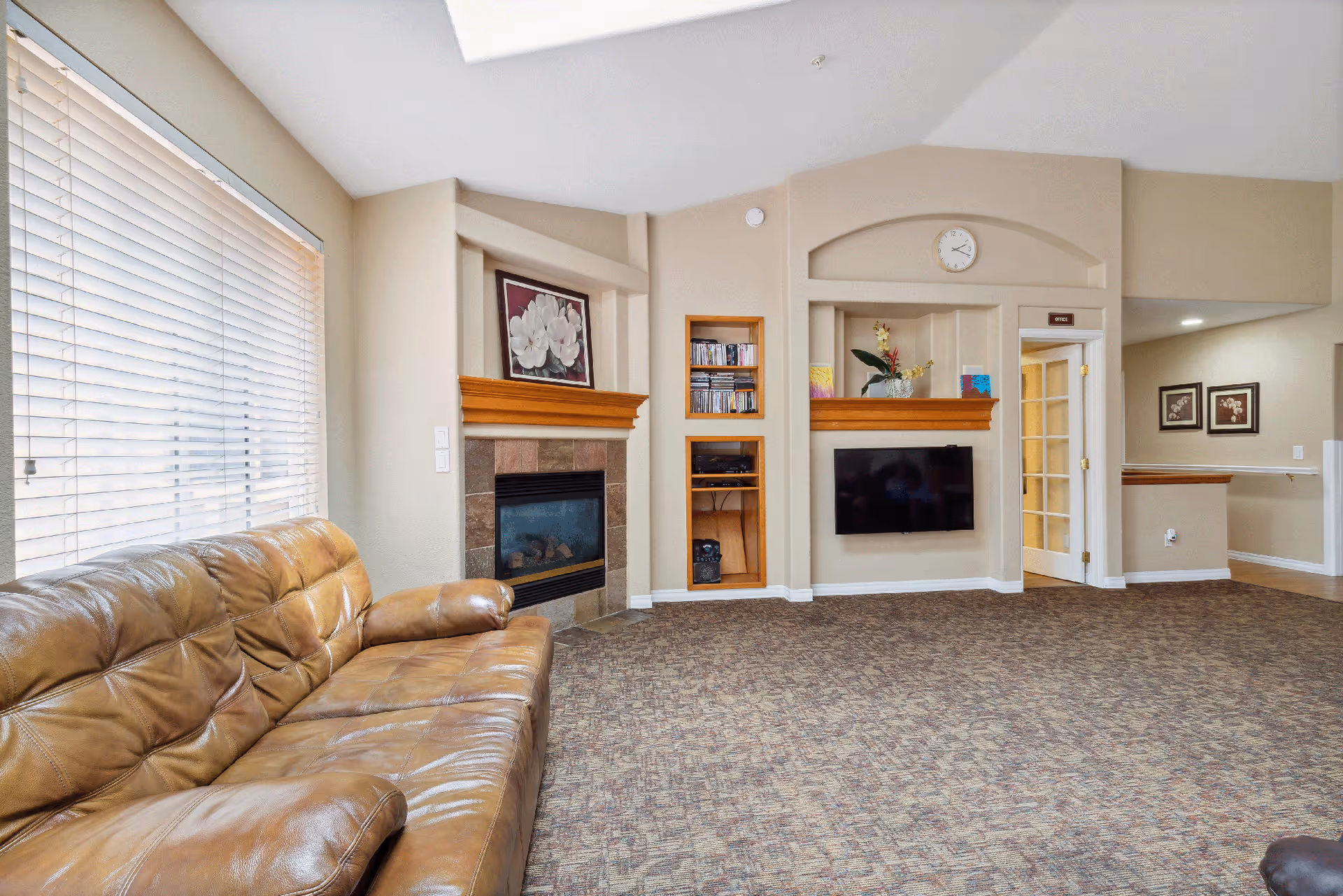 A cozy living room with a large brown leather sofa on the left, a window with white blinds, a fireplace with a floral painting above it, built-in shelves holding books and decorative items, a wall-mounted flat-screen TV, and a clock above an arched wall niche. The room has beige walls and patterned carpet flooring.