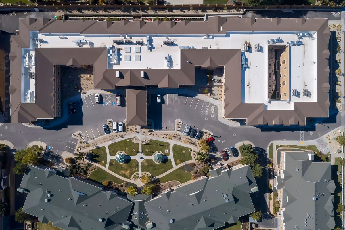 Aerial view of Crescent Senior Living facility showing the building rooftops, parking lots with several cars, landscaped garden area with pathways and gazebos, and surrounding roads.