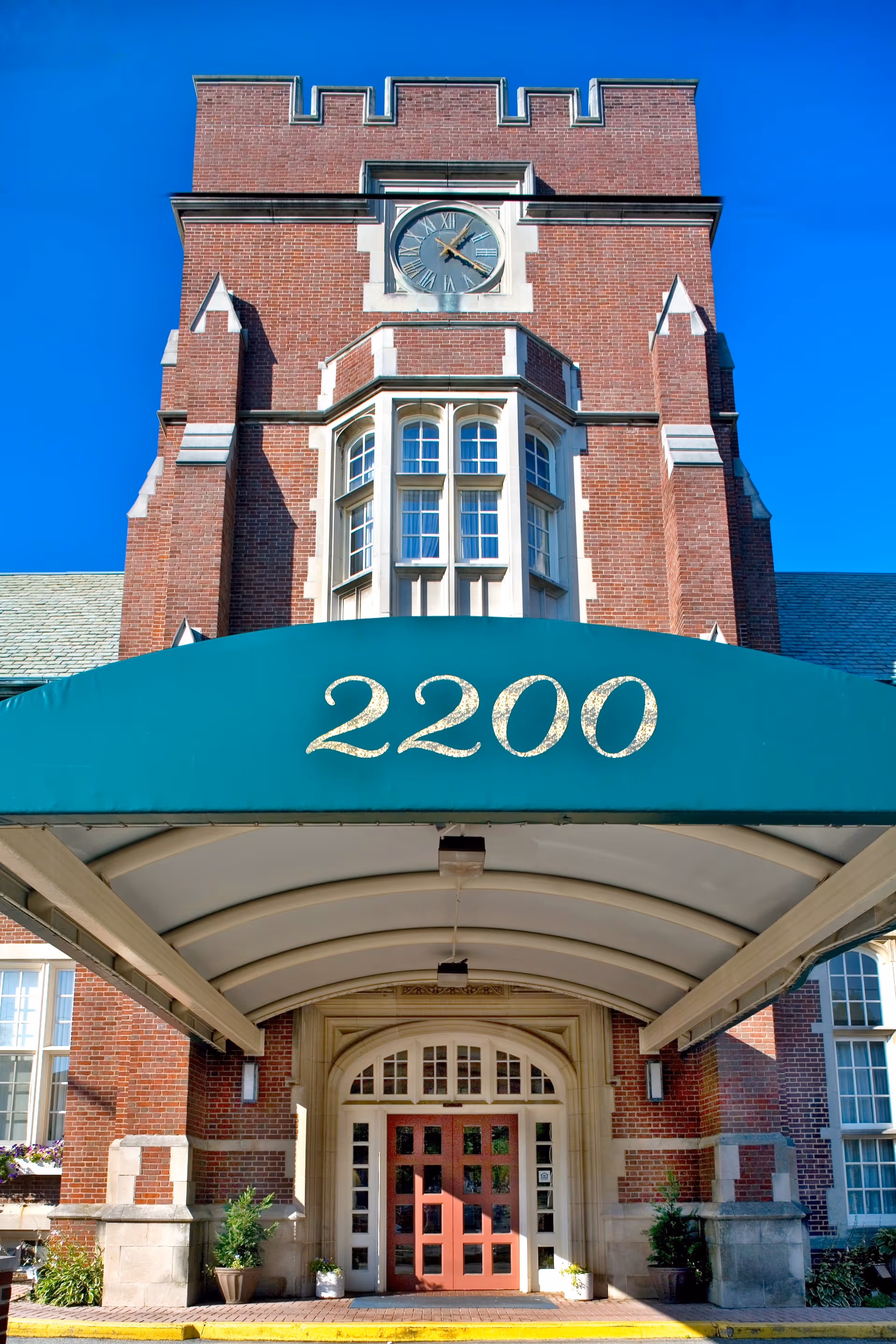 Entrance of a brick building with a large green awning displaying the number 2200. The building features a clock above a bay window and an arched doorway with glass panels. There are potted plants on either side of the entrance.