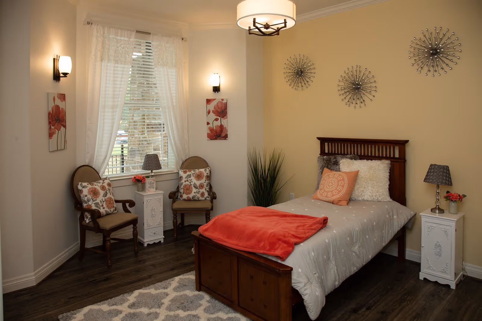 Cozy decorated bedroom with a single wooden bed, bedside tables, two chairs by a window, and wall art.
