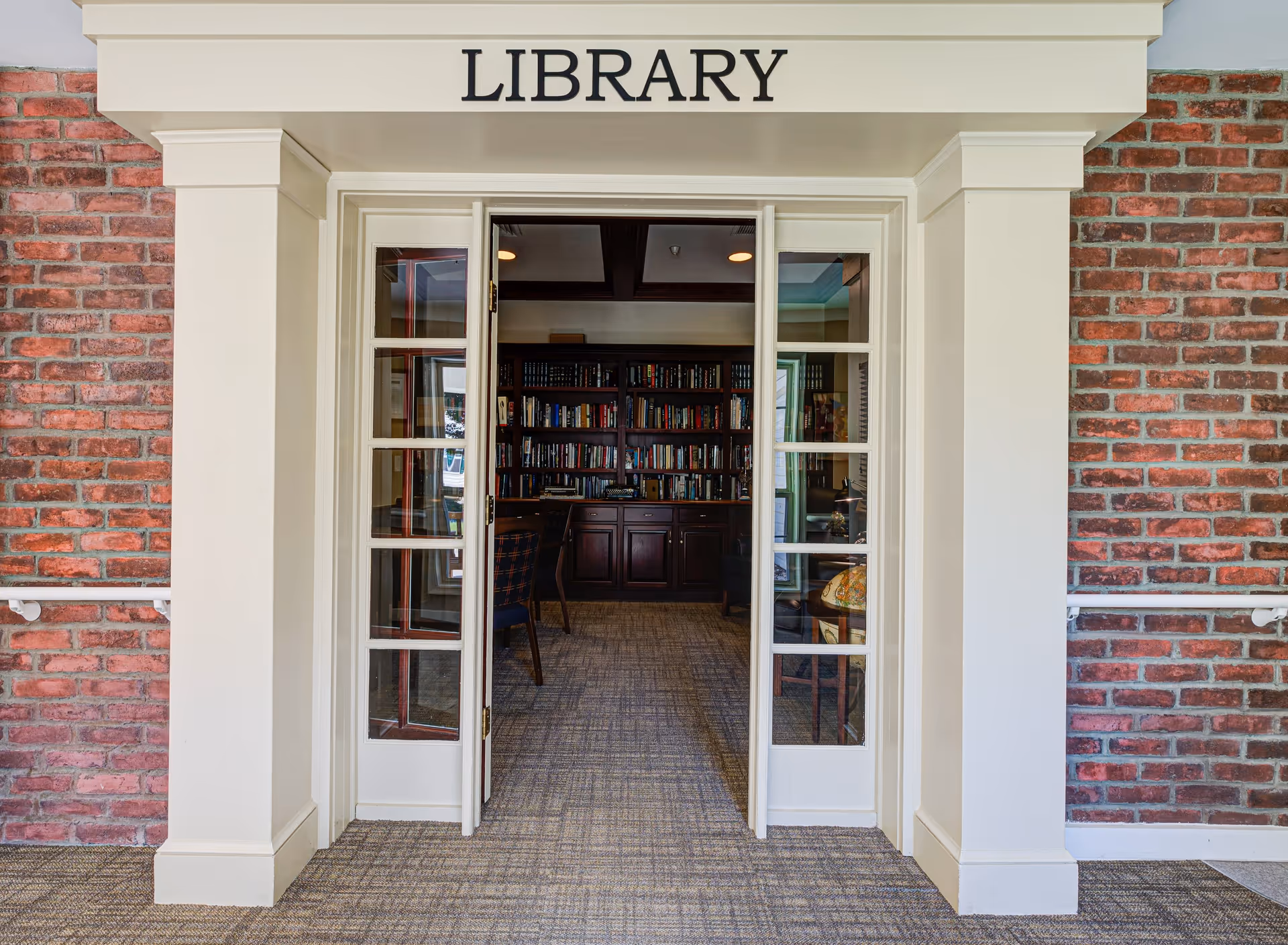 Entrance to a library room with white framed double glass doors open, revealing bookshelves filled with books and a few chairs inside. The entrance is surrounded by red brick walls and has a sign above the door that reads 'LIBRARY'.