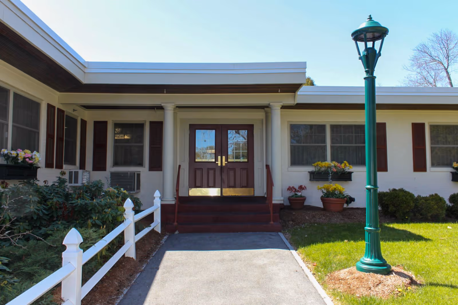 Entrance to a single-story building with double wooden doors, white columns, and windows with brown shutters. There is a white fence along a walkway leading to the entrance, flower pots with colorful flowers, green bushes, and a green lamppost on a grassy area.