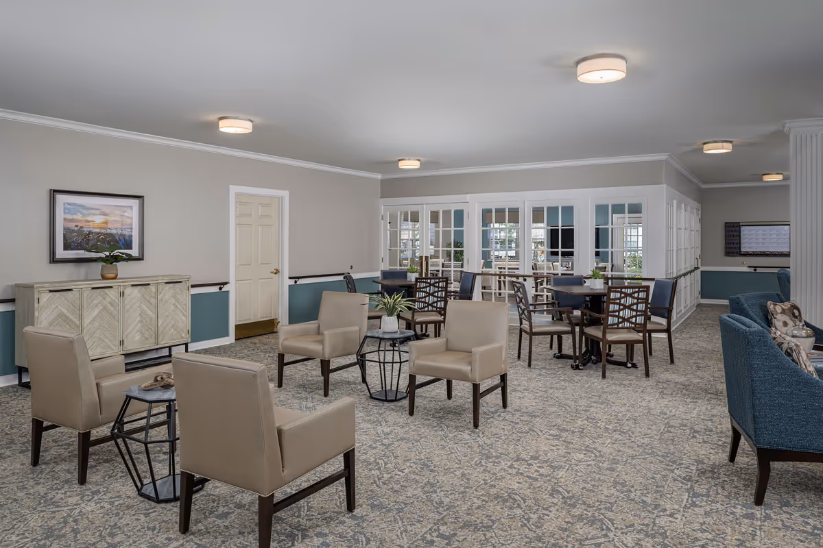 A spacious and well-lit common area in a senior living facility with beige armchairs arranged around small tables, additional dining tables with chairs, a blue couch with patterned pillows, and a sideboard with a plant and framed artwork on the wall. The room has light gray walls, a patterned carpet, and multiple ceiling lights.