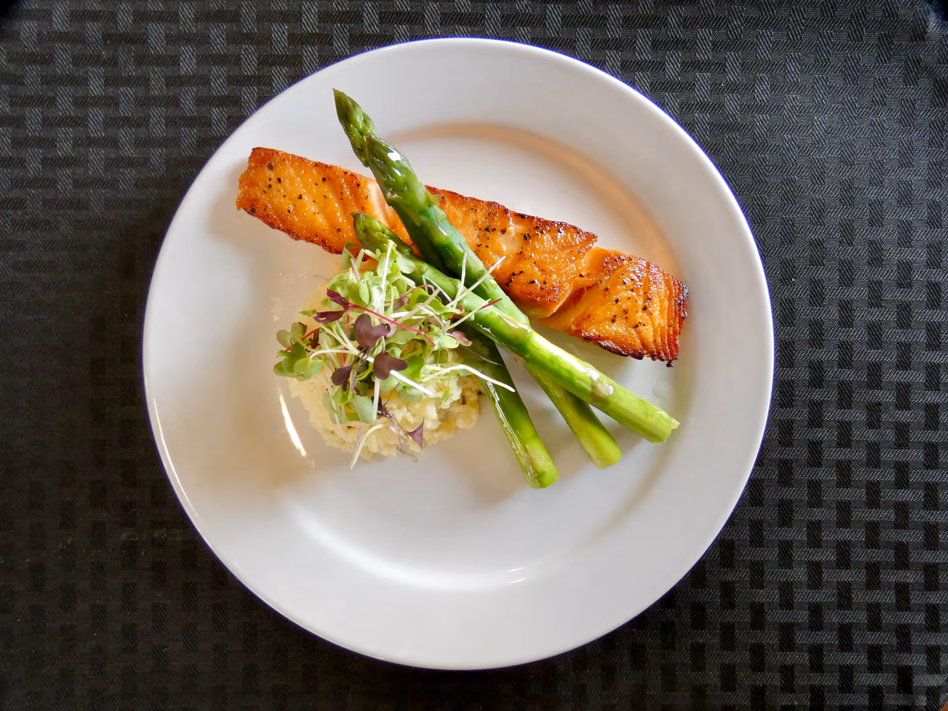 A white plate with a serving of grilled salmon, three asparagus spears, and a small mound of mashed potatoes topped with microgreens, placed on a dark textured placemat.