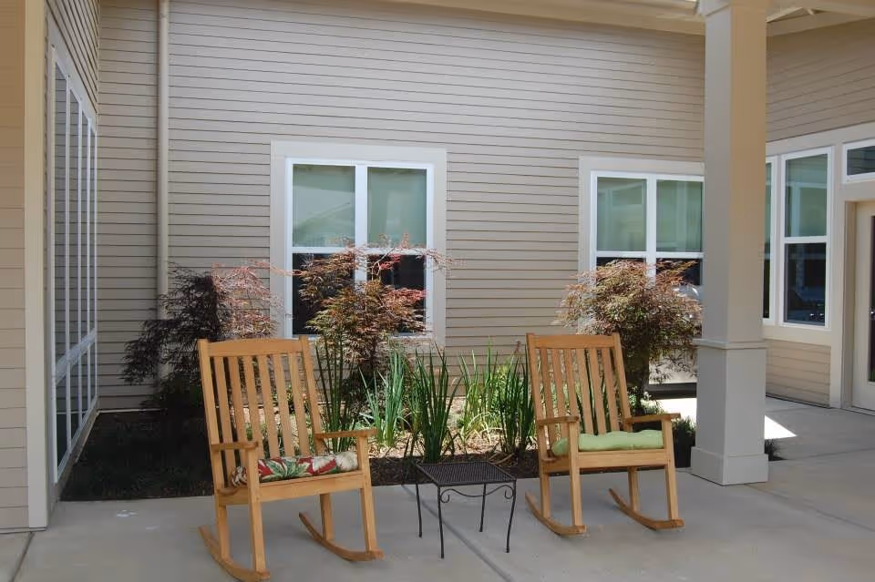 Outdoor patio area with two wooden rocking chairs with cushions and a small metal table between them. The area is surrounded by beige siding walls with windows and some plants and shrubs in a garden bed.