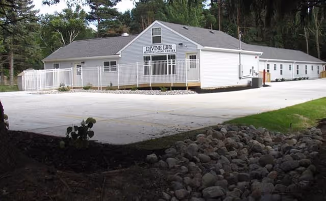 Exterior view of a single-story white building with a sign that reads 'Divine Life Assisted Living Center'. The building is surrounded by a paved area, a white fence, and some landscaping with rocks and grass. Trees are visible in the background.