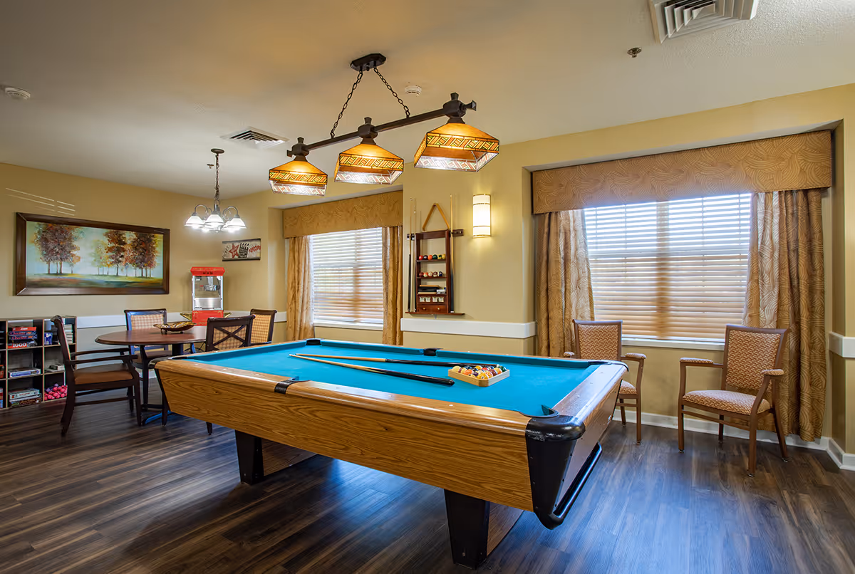 A well-lit recreation room with a pool table, seating, windows with blinds, and a games area with a popcorn machine.