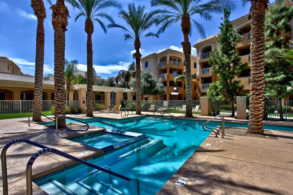 Outdoor swimming pool area at The Heritage Tradition facility, surrounded by tall palm trees and multi-story residential buildings under a clear blue sky. The pool area includes a hot tub, metal handrails, and lounge chairs with a fenced perimeter.