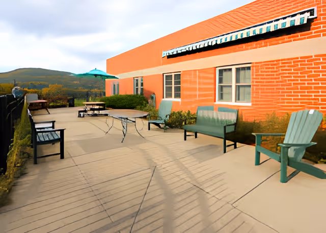 Outdoor patio area with several benches, chairs, and tables next to a red brick building. There is a green umbrella providing shade over one table, and a scenic view of hills and trees in the background under a partly cloudy sky.