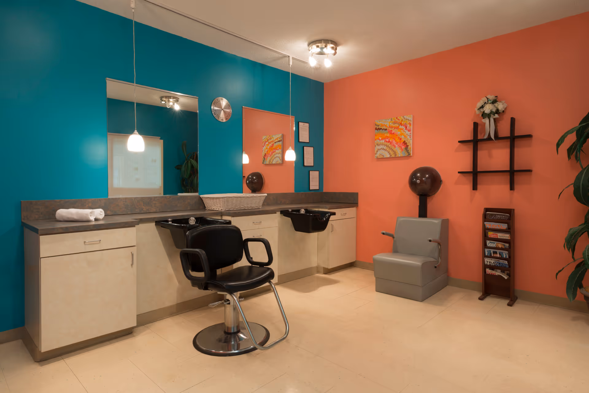 Interior view of a hair salon area in a senior living facility with teal and coral walls. The room features two black salon chairs in front of sinks, a countertop with cabinets, a wall-mounted hair dryer, a magazine rack, a decorative wall shelf with flowers, and colorful artwork on the walls.