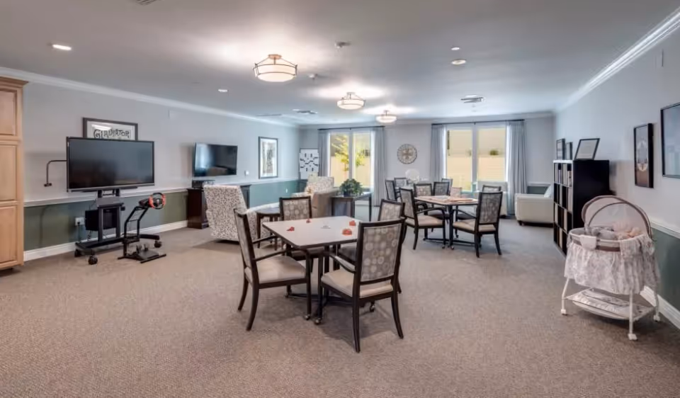 A spacious common area in a senior living facility with multiple tables and chairs arranged for group activities or dining. The room features two flat-screen TVs mounted on the walls, comfortable armchairs, large windows with curtains allowing natural light, and a bassinet on the right side. The walls are painted in a two-tone style with white on top and green on the bottom, and the ceiling has several light fixtures.